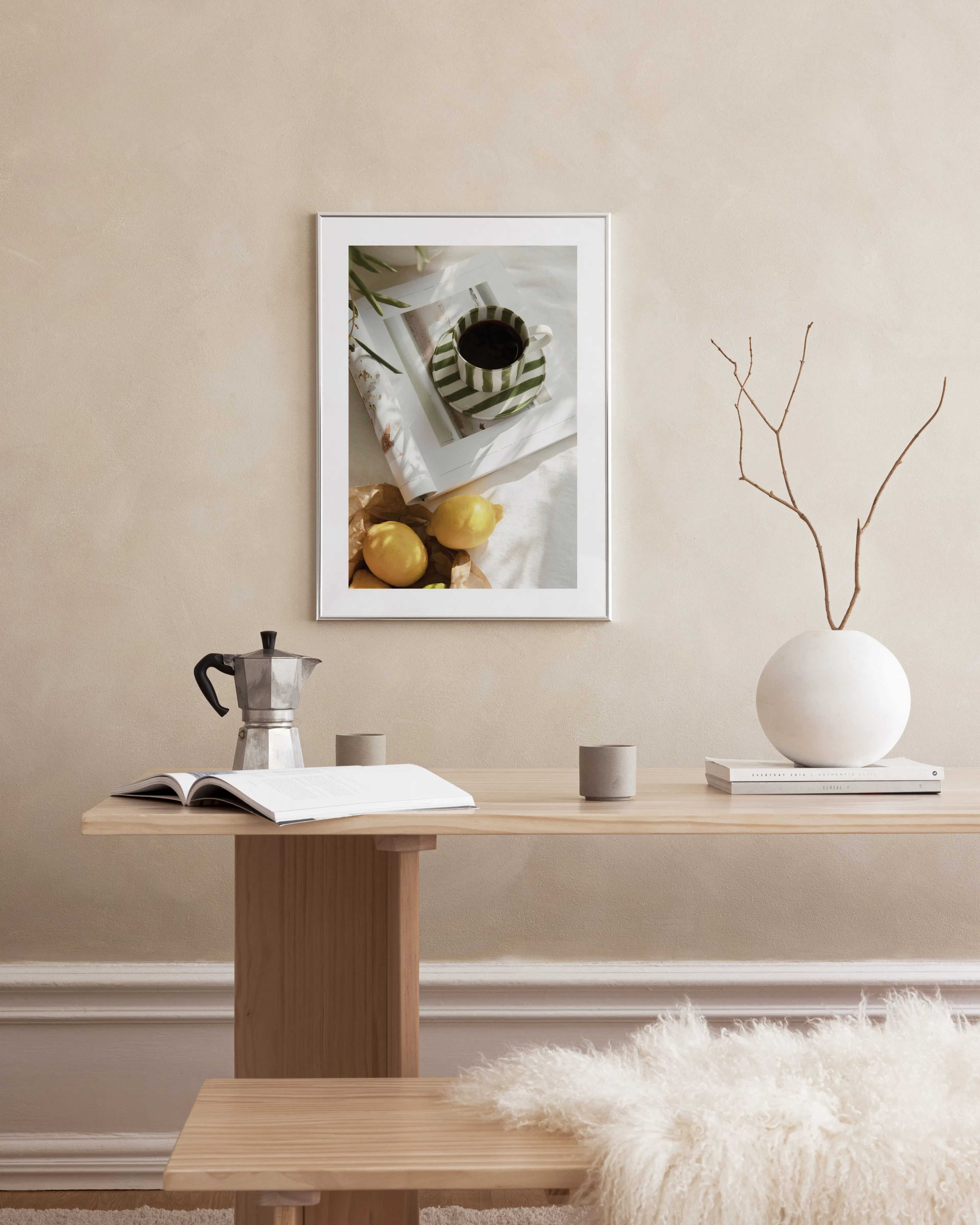 Affiche avec tasse à café rayée verte, citrons et magazine, accrochée au-dessus dune table en bois avec cafetière et vase.