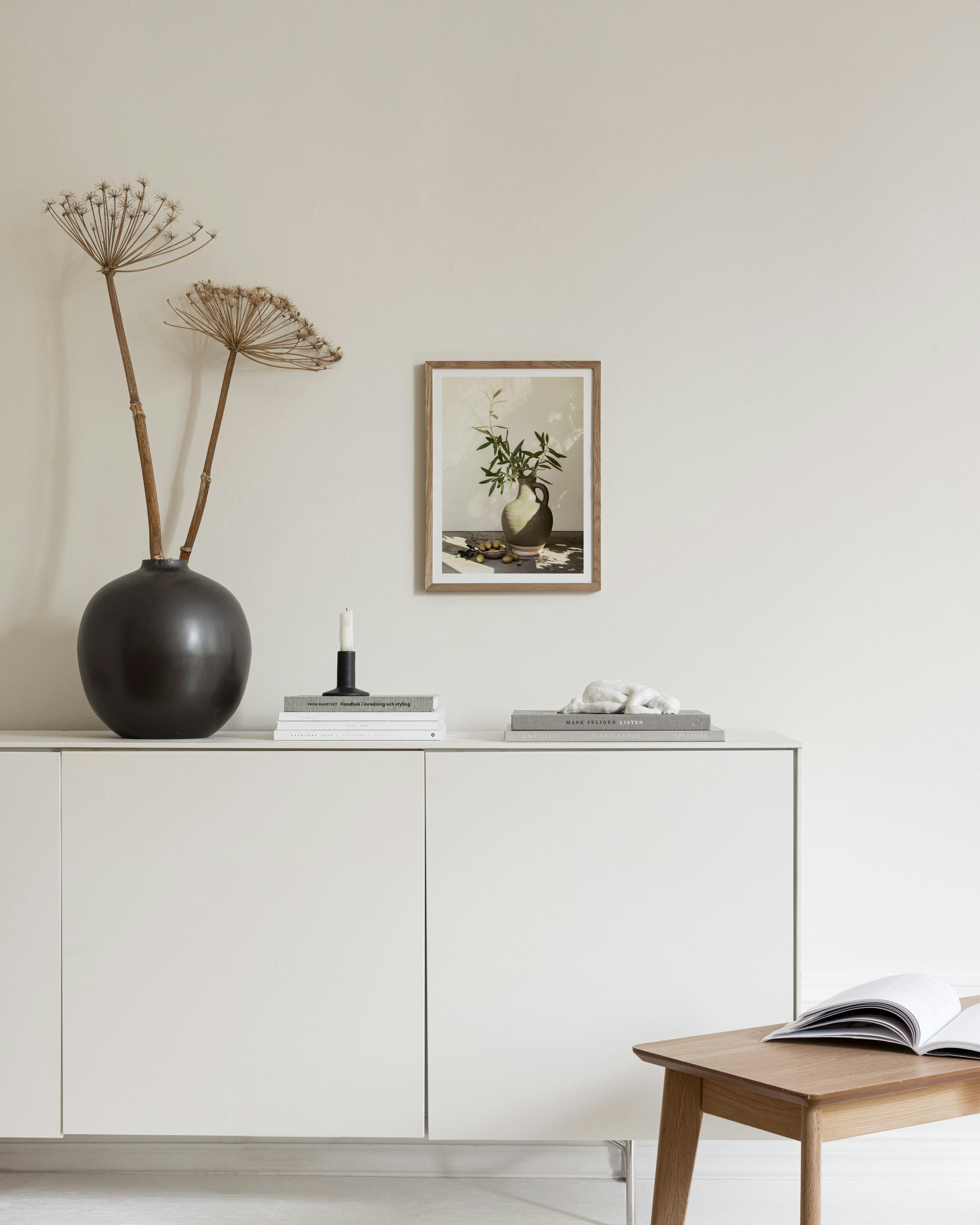 Poster con rami dolivo in vaso di ceramica e olive, appeso sopra una credenza bianca in un salotto.
