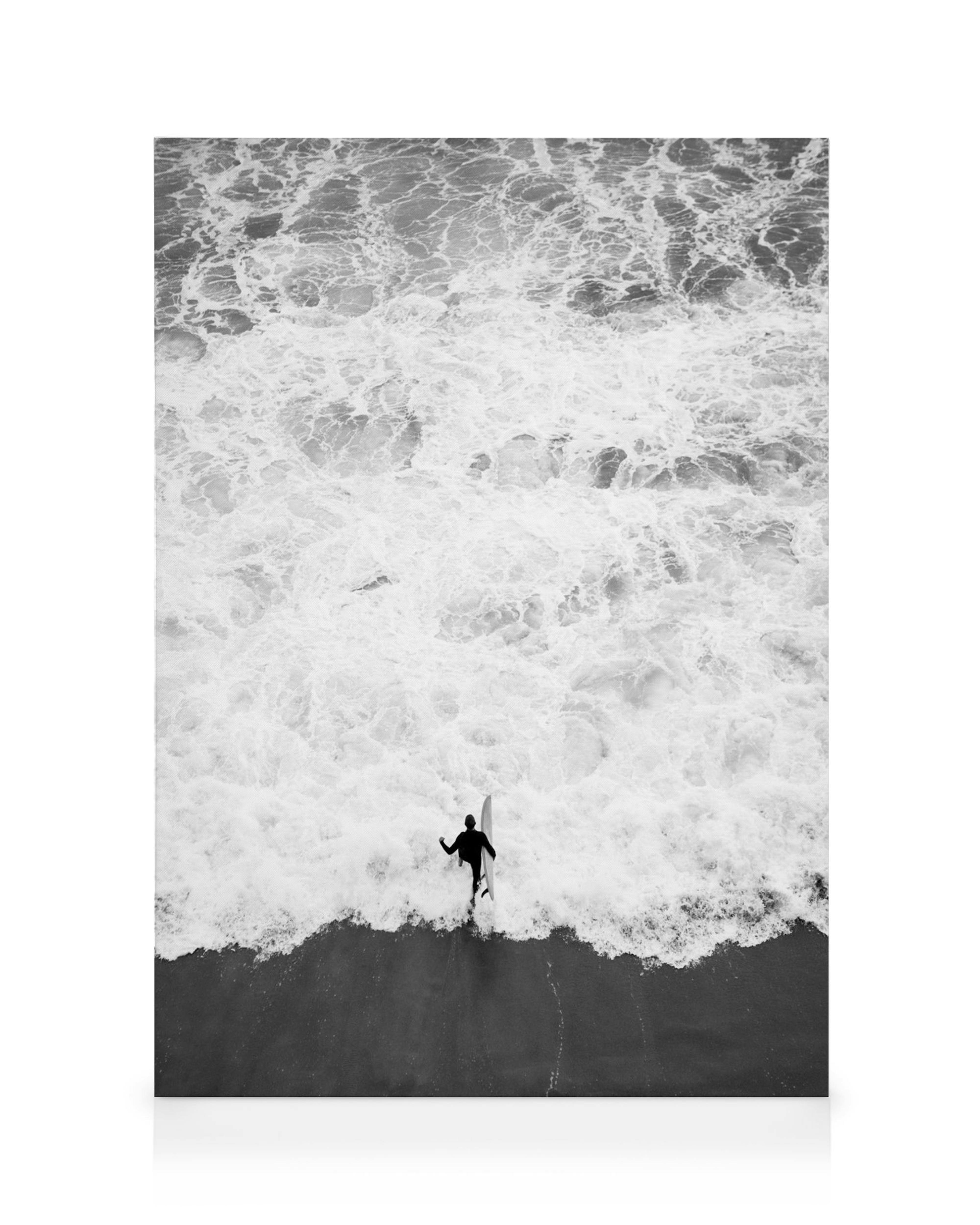A black and white canvas print of a lone surfer walking into the ocean with a surfboard, surrounded by foamy waves.