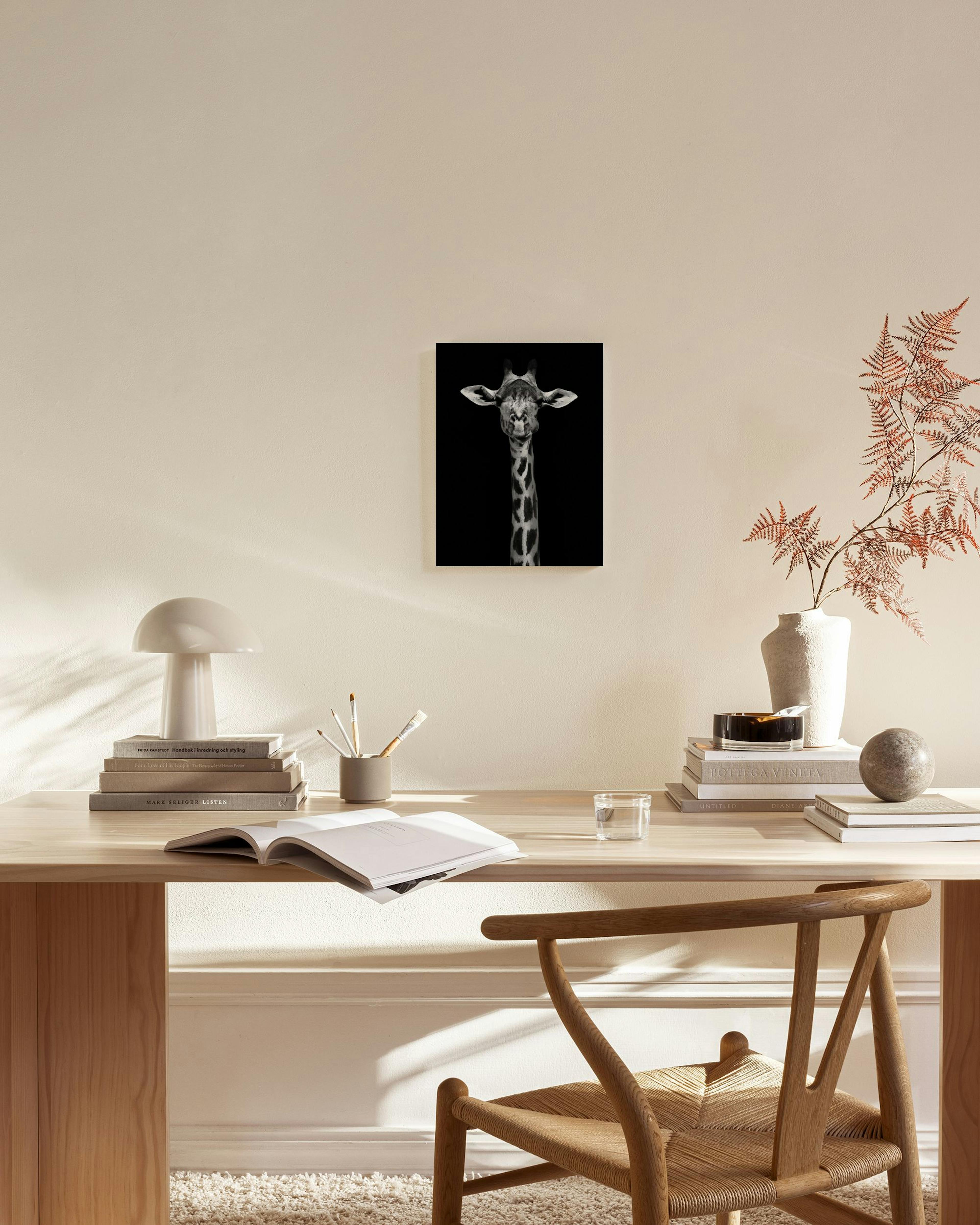 Lærred med en sort-hvid afbildning af en giraf mod sort baggrund, hængende i en stue over et træmøbel.