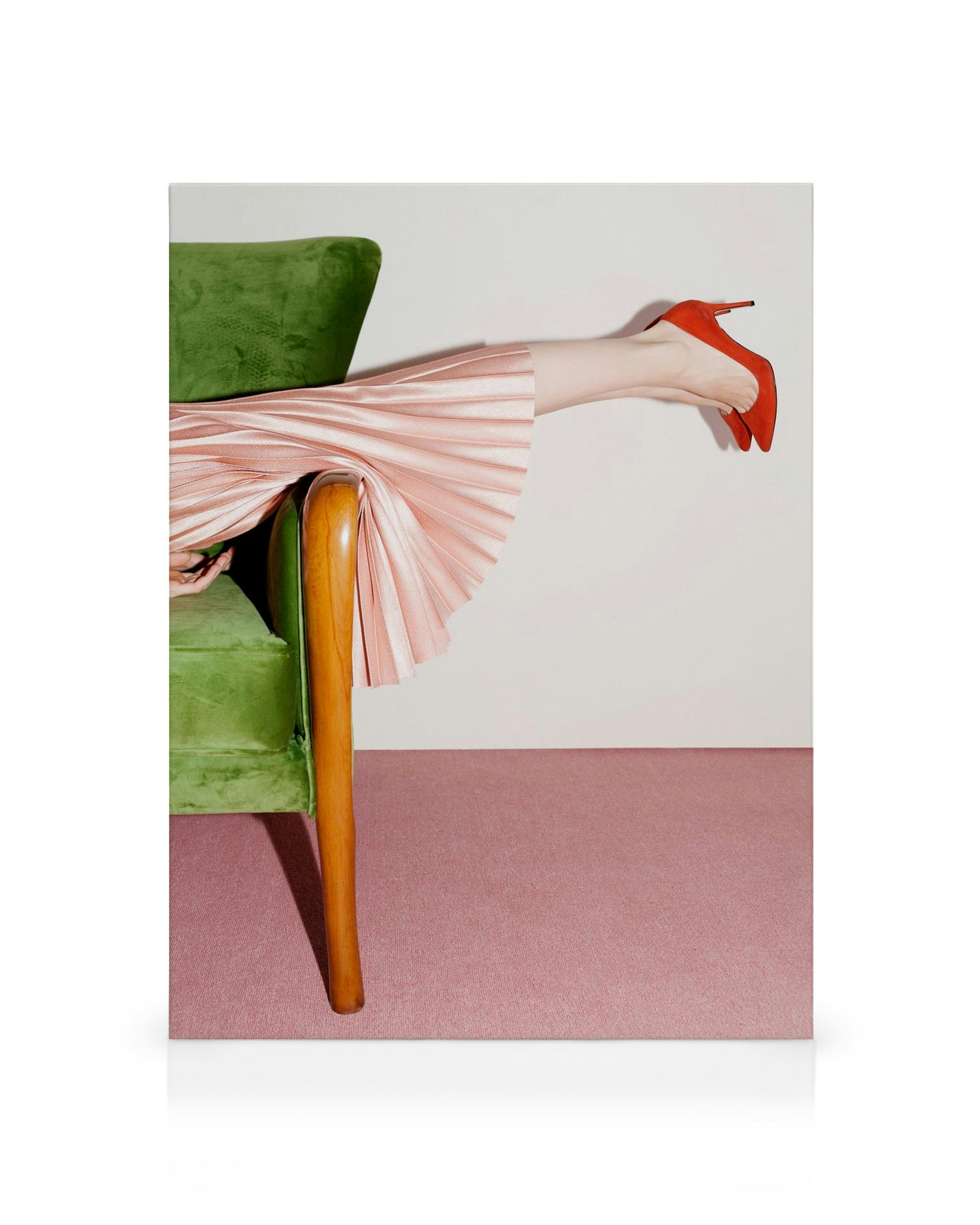 A person reclines on a green armchair with a pleated pink skirt and red high heels, legs extended against a white wall and pink 