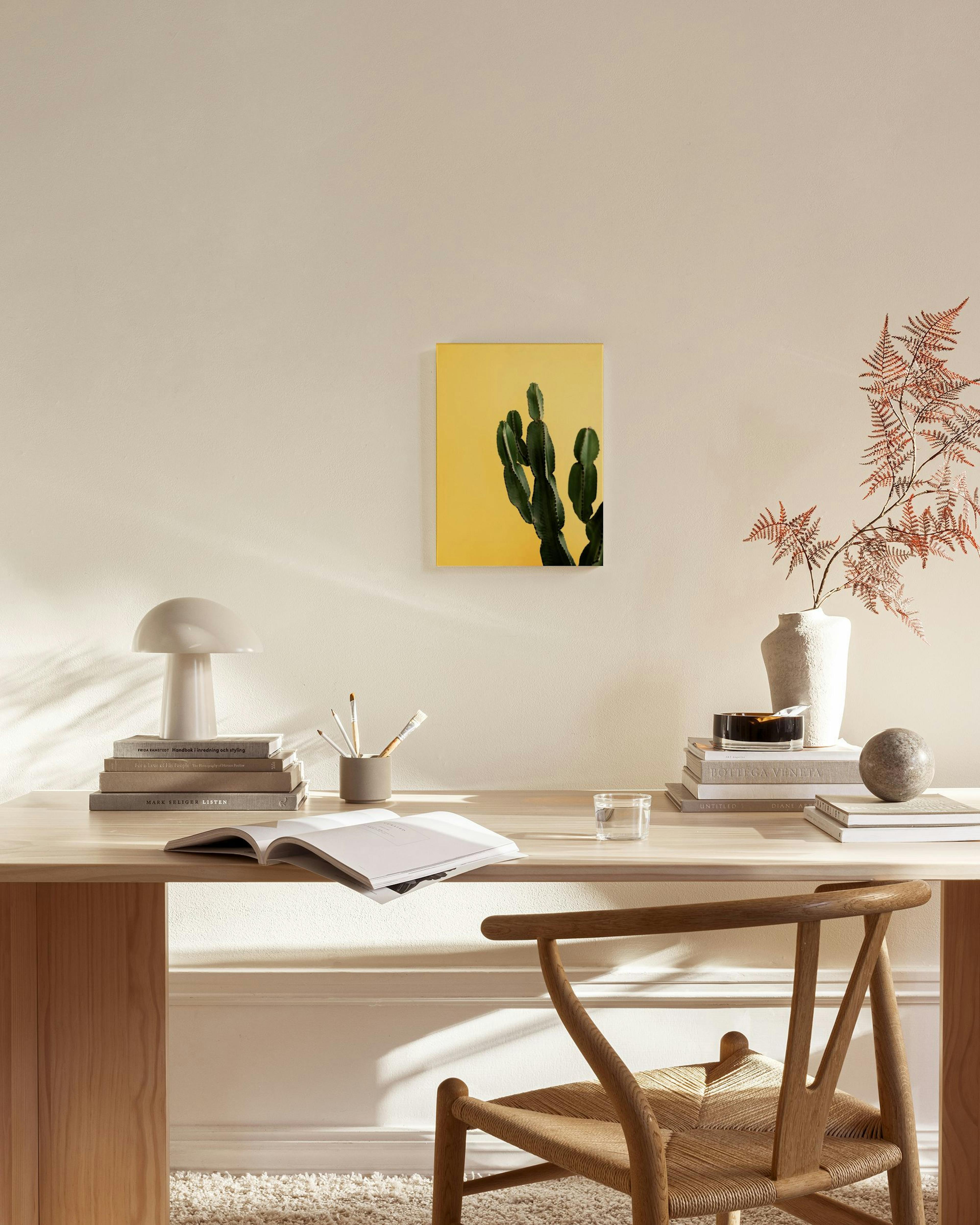 Obraz kaktusu na žlutém pozadí visí nad dřevěnou komodou v moderním obývacím pokoji s křeslem a skleněným stolkem.