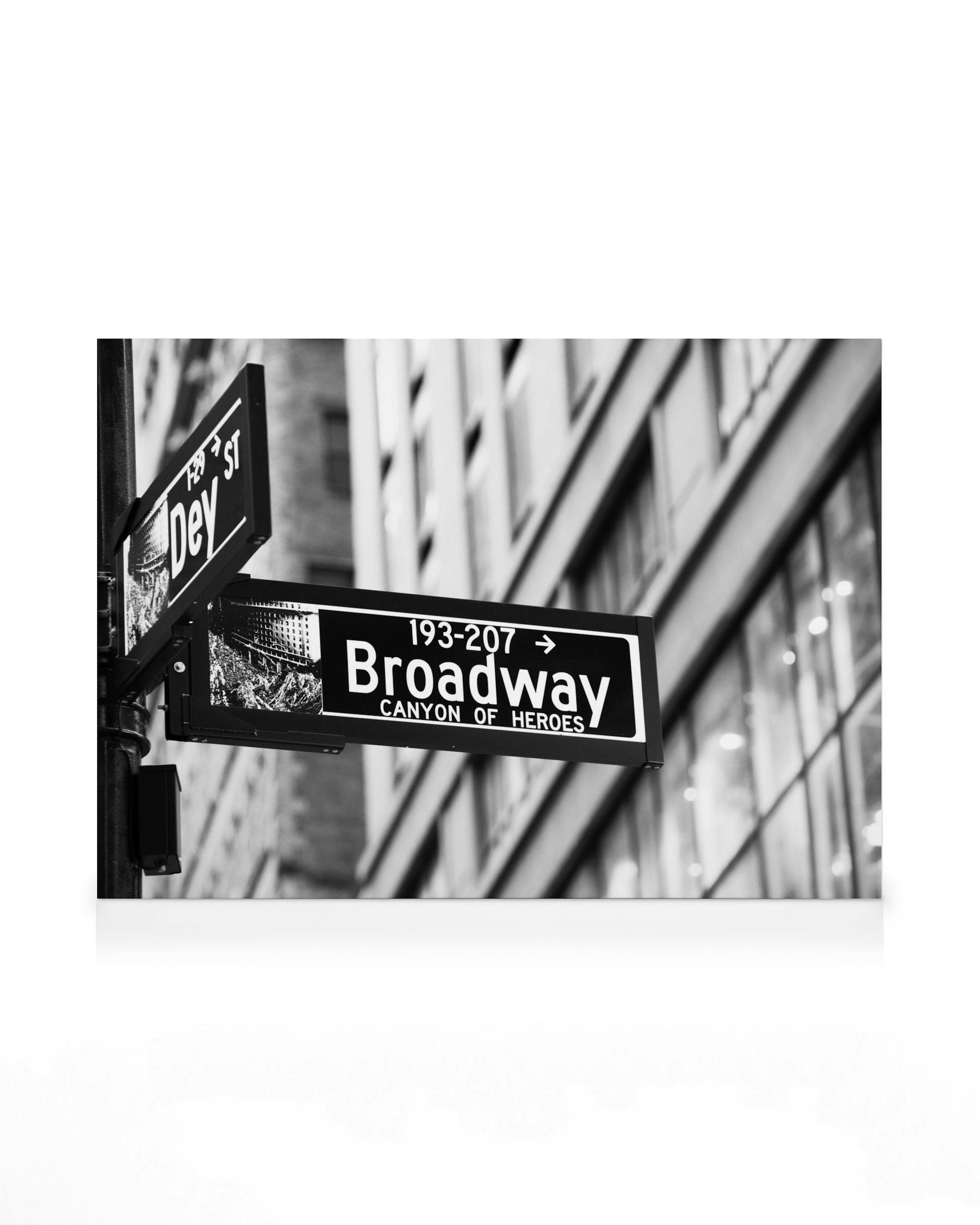 Black and white Broadway street sign in New York City, featuring CANYON OF HEROES text, with blurry buildings in the background.