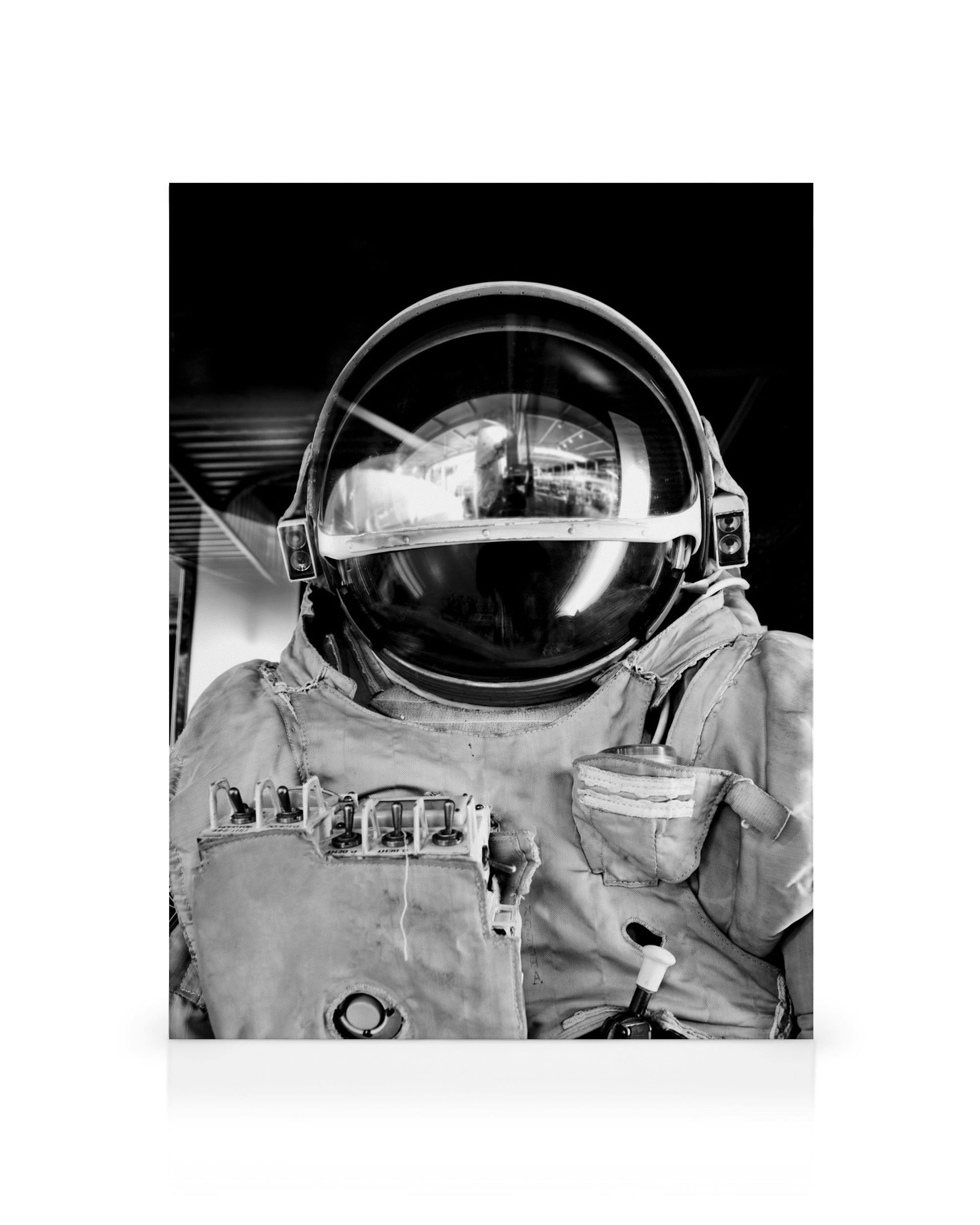 Čiernobiely obraz astronauta v skafandri s prilbou, odrážajúcou okolie, detaily ovládacieho panela.