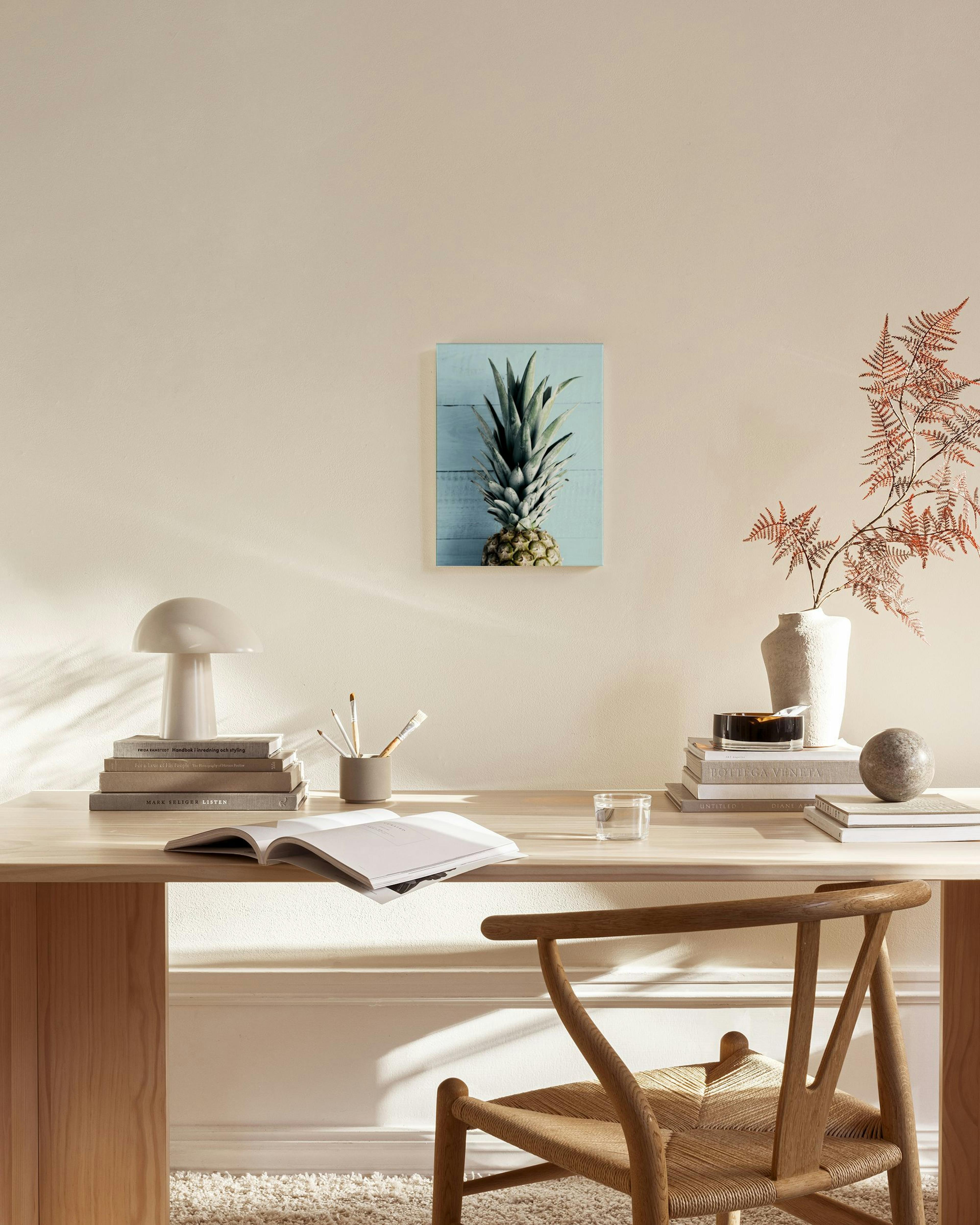 Lærred med ananas på lyseblå baggrund, hængende over et lavt træmøbel i en stue.