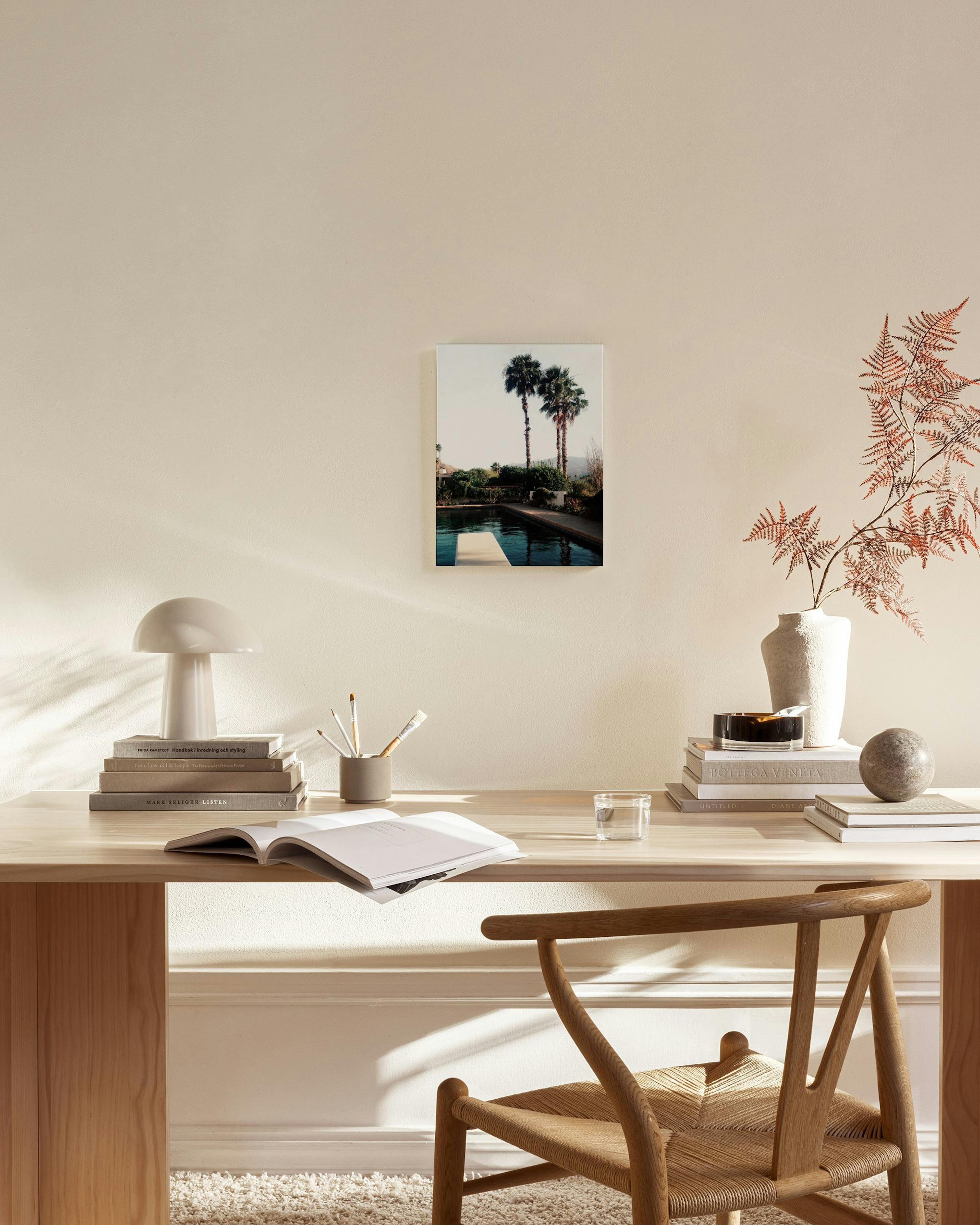 A canvas print of two palm trees by a swimming pool and diving board, hanging in a bright, minimalist living room.