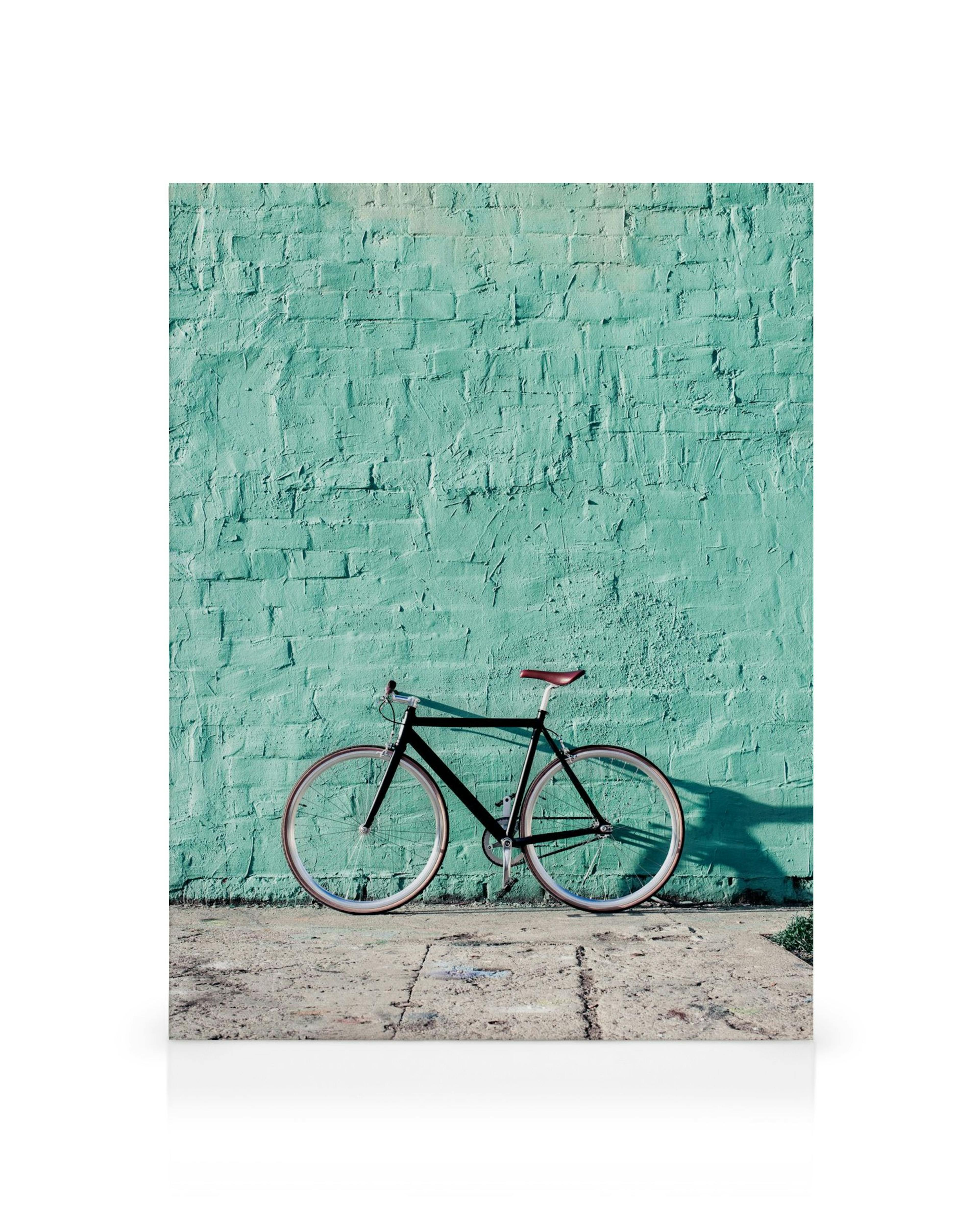 Bicicleta negra con ruedas marrones y asiento rojo, apoyada en una pared turquesa texturizada, ideal para decoración de pared.