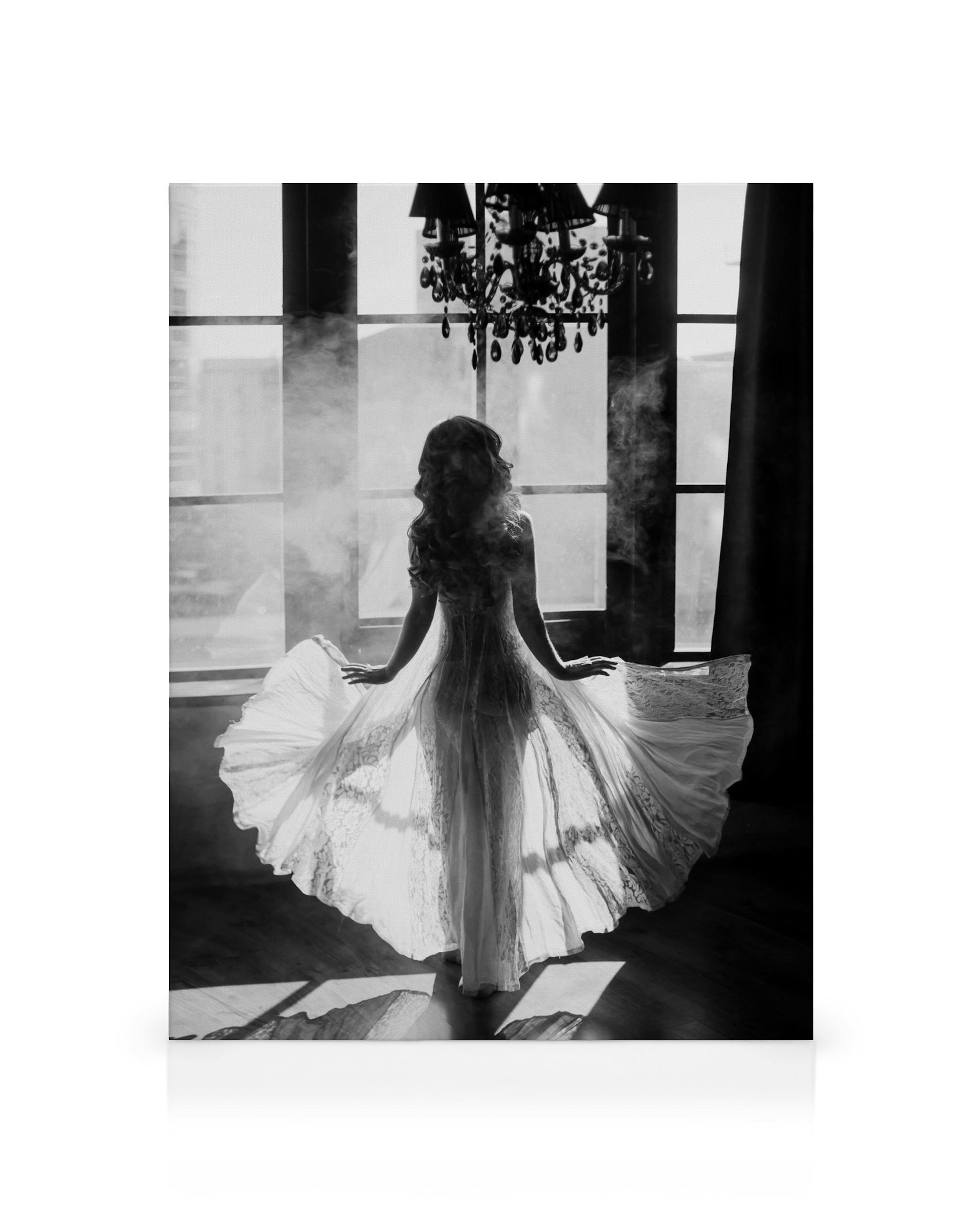 Black and white canvas print of a woman in a flowing white dress, back to the viewer, standing by a window with a chandelier.
