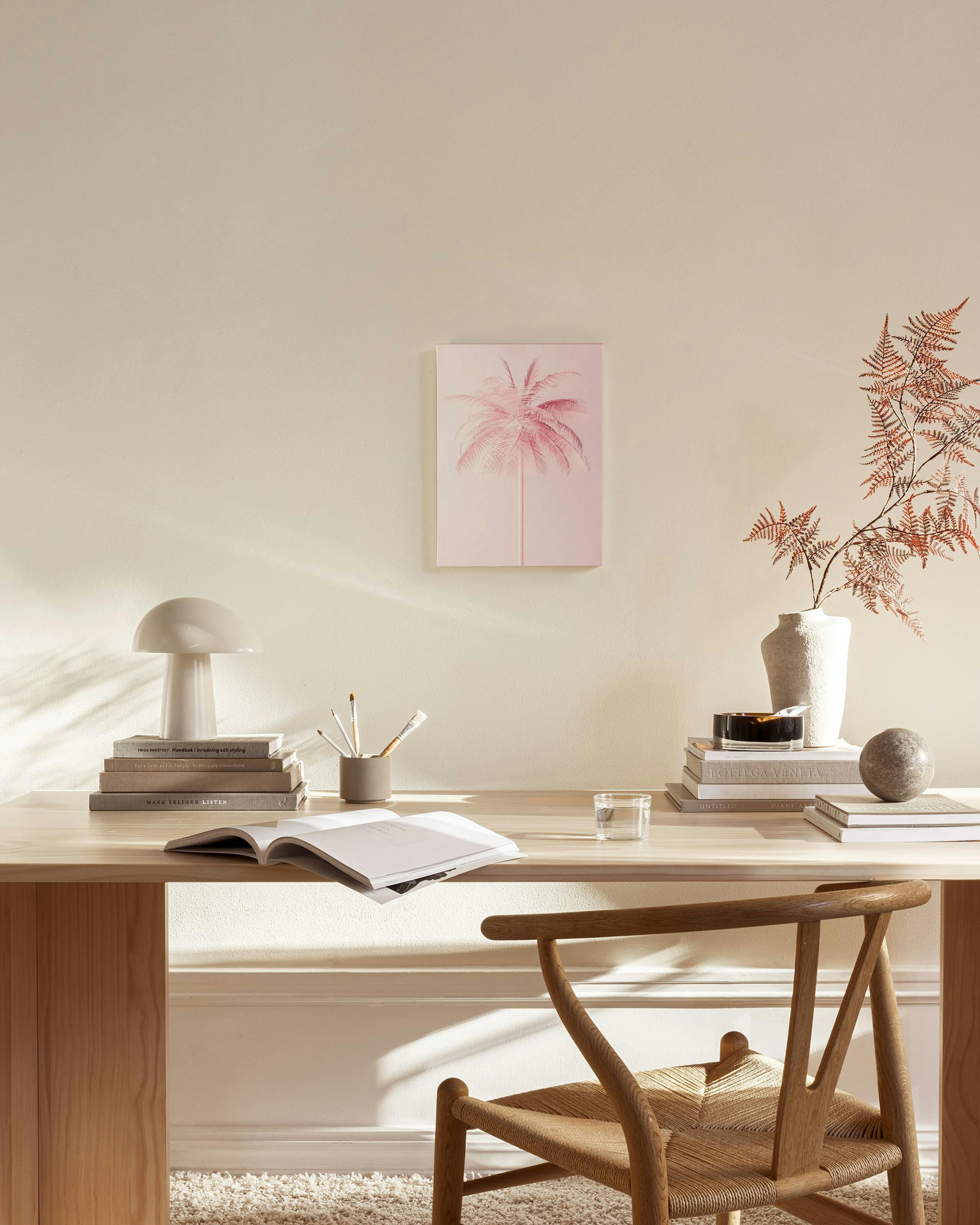 A pink palm tree print hangs in a minimalist living room with a wooden chair and glass coffee table.