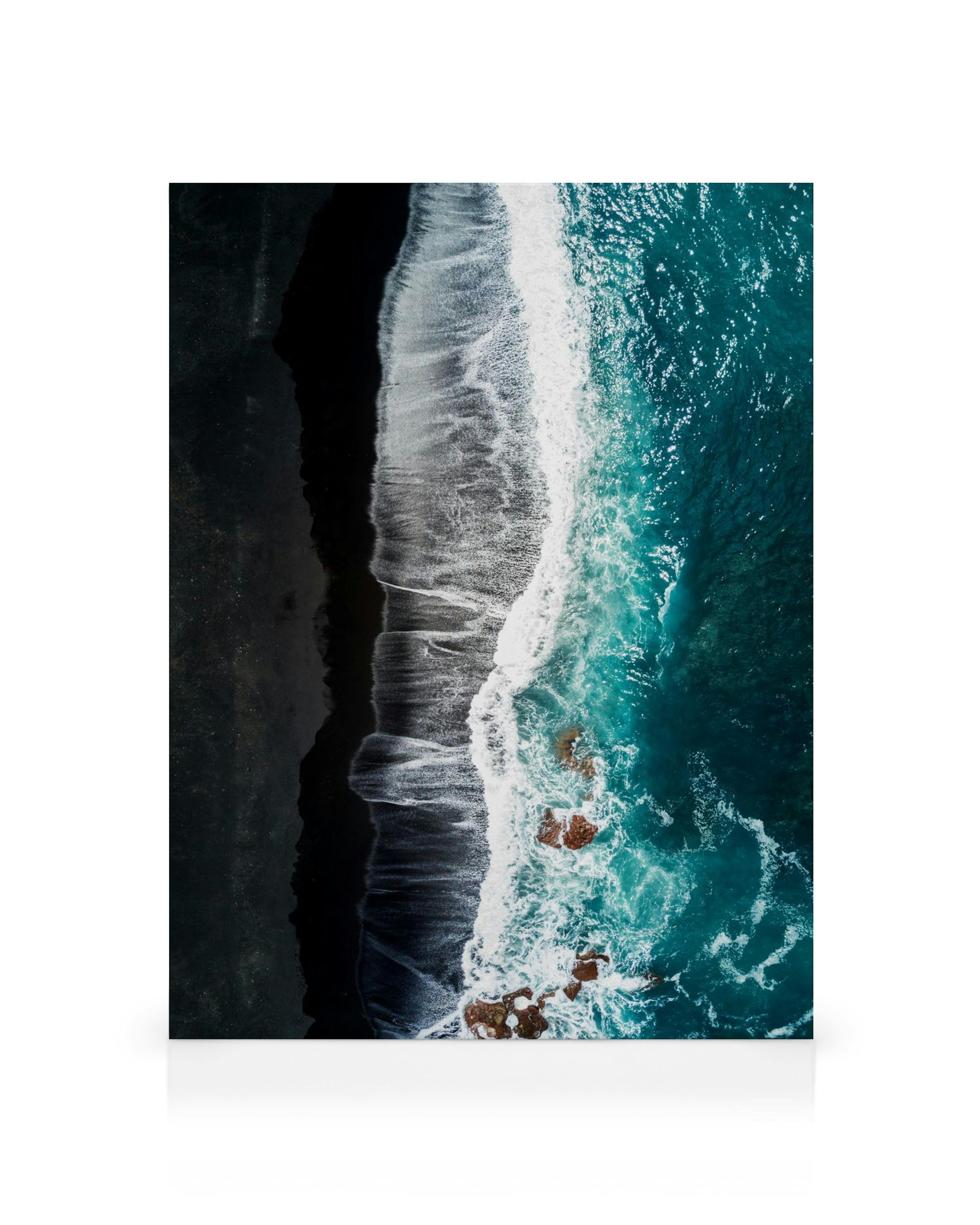 Flygfoto av svart sandstrand med turkosblå vågor som slår mot strandlinjen, exponerade klippor i havet.