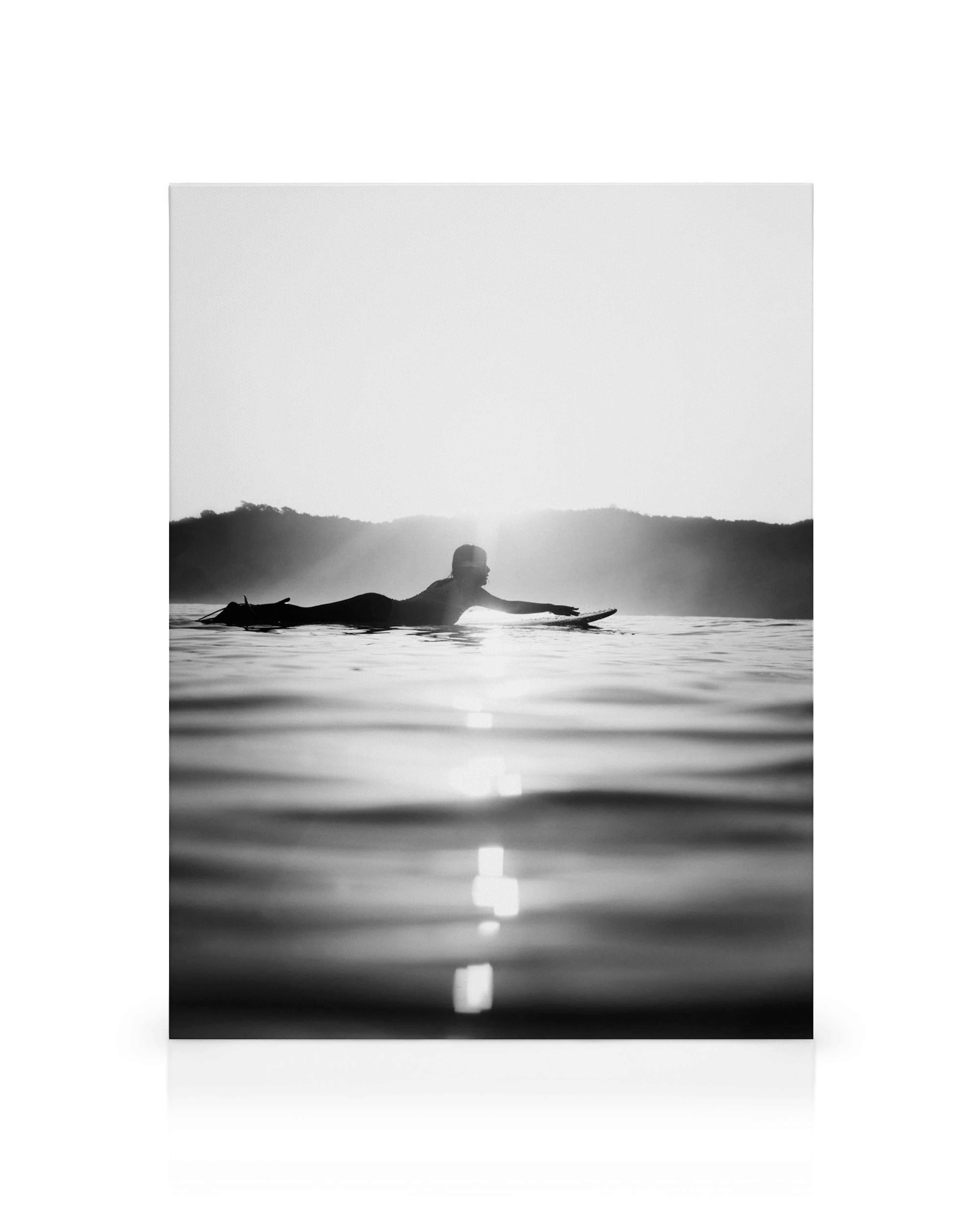 Svartvit canvastavla med en person som paddlar på en surfbräda i ett lugnt vattenlandskap med berg i bakgrunden.