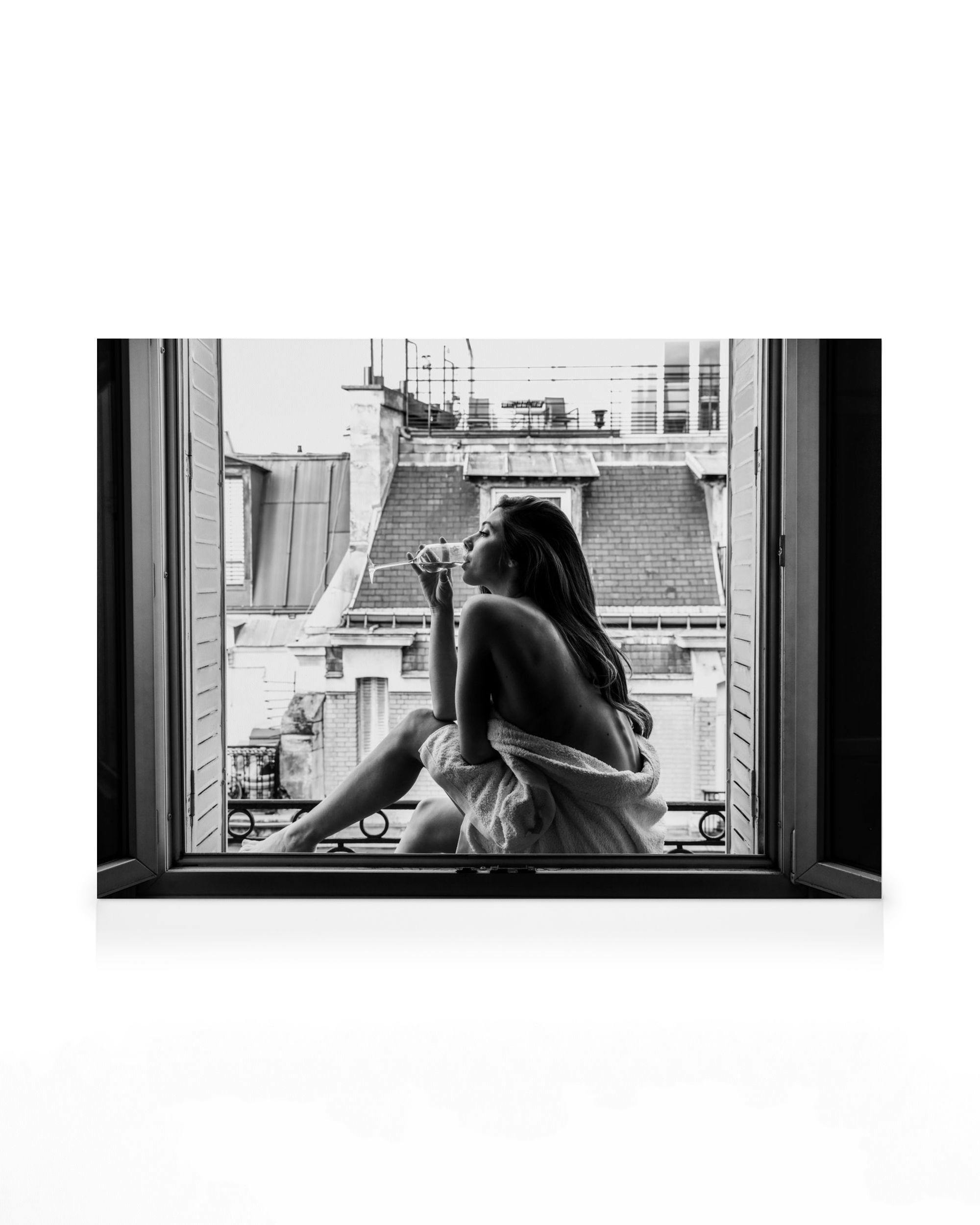 Black and white photograph of a woman in a robe drinking wine on a balcony overlooking Parisian rooftops.
