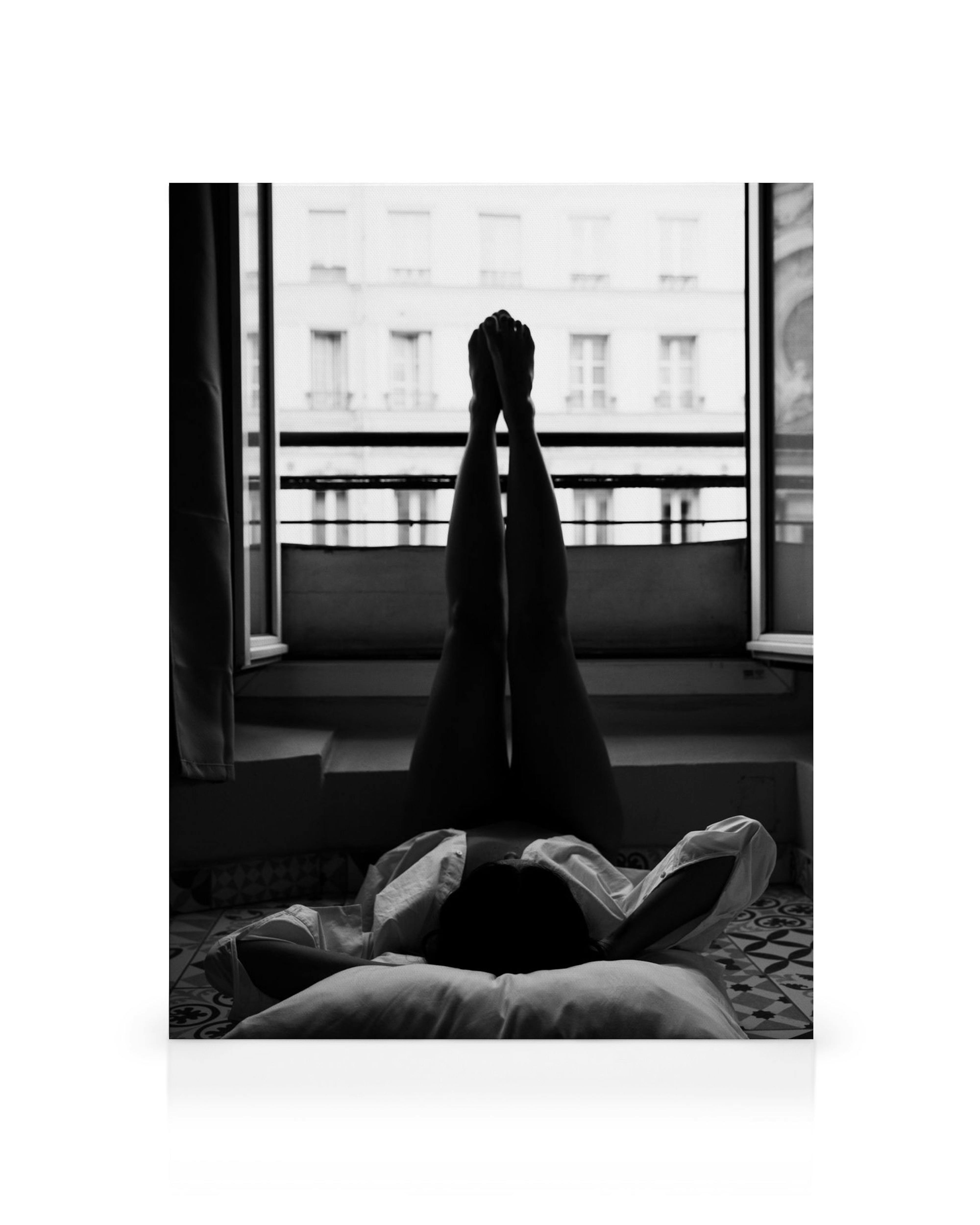Svart-hvitt lerret av person som ligger på gulvet med bena opp ved et åpent vindu med utsikt over Paris.