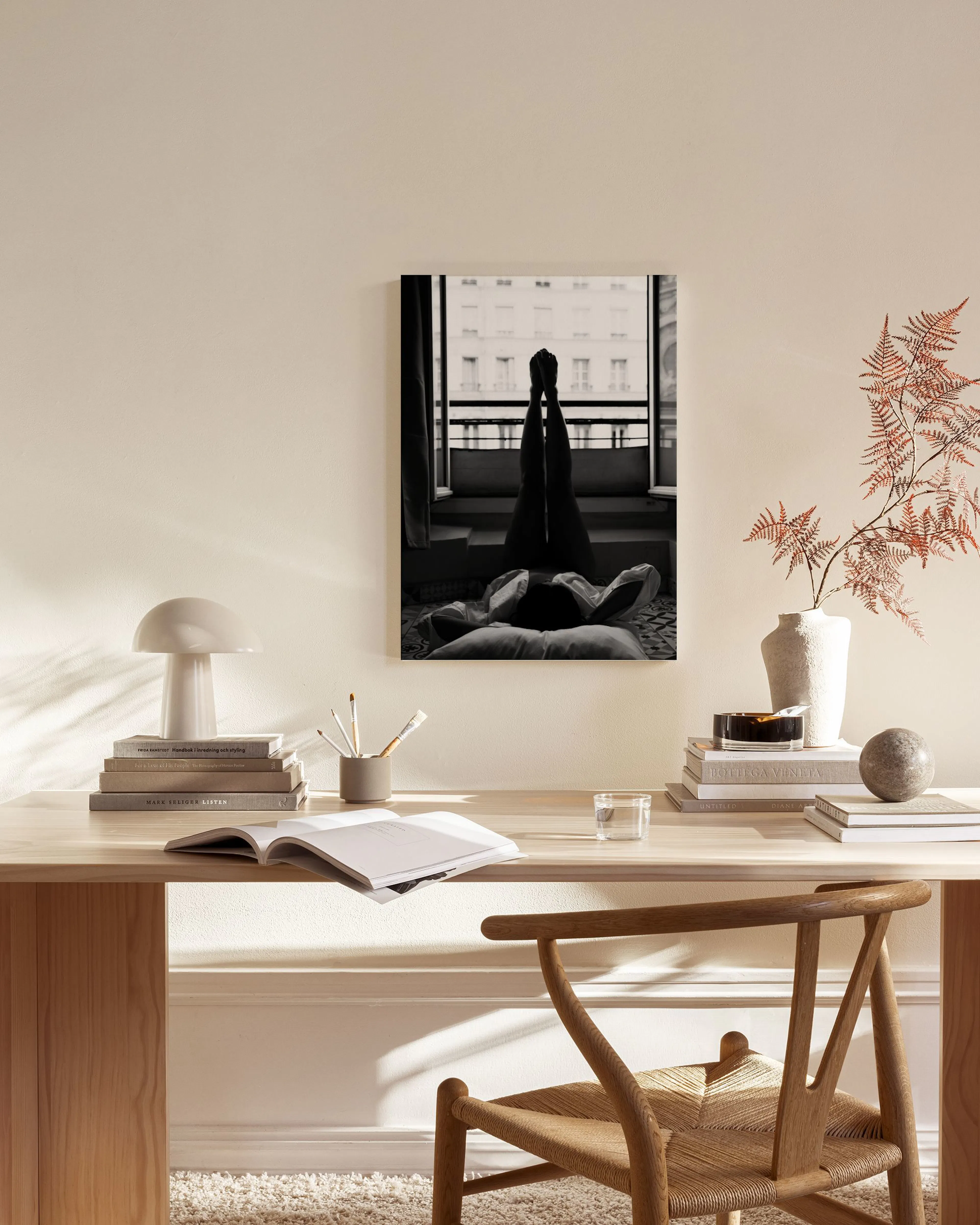 A black and white canvas print featuring legs resting on a bed by a large window, hanging in a bright, minimalist living room.