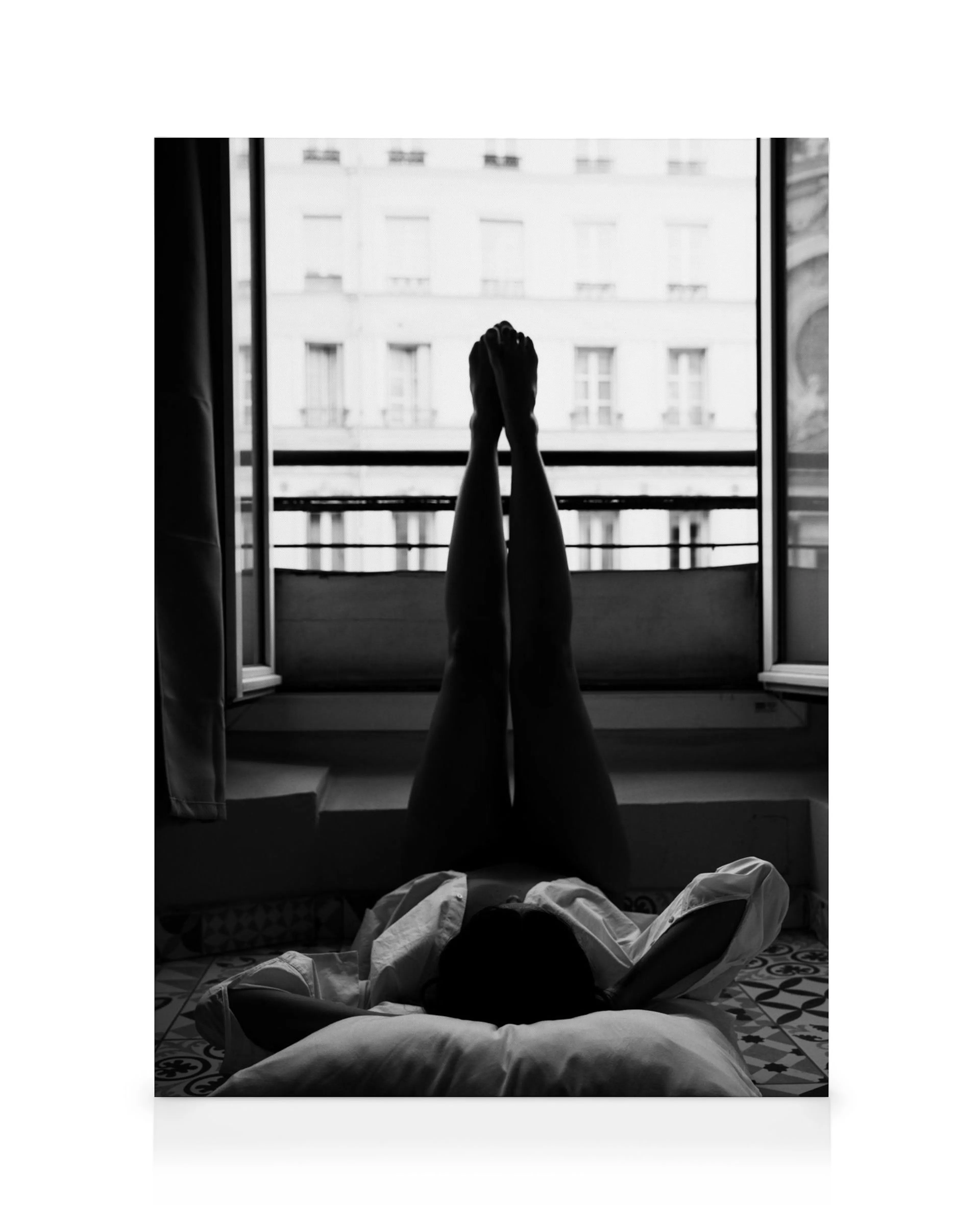 Black and white Paris window view from a bed, with a persons legs silhouetted against the urban skyline.