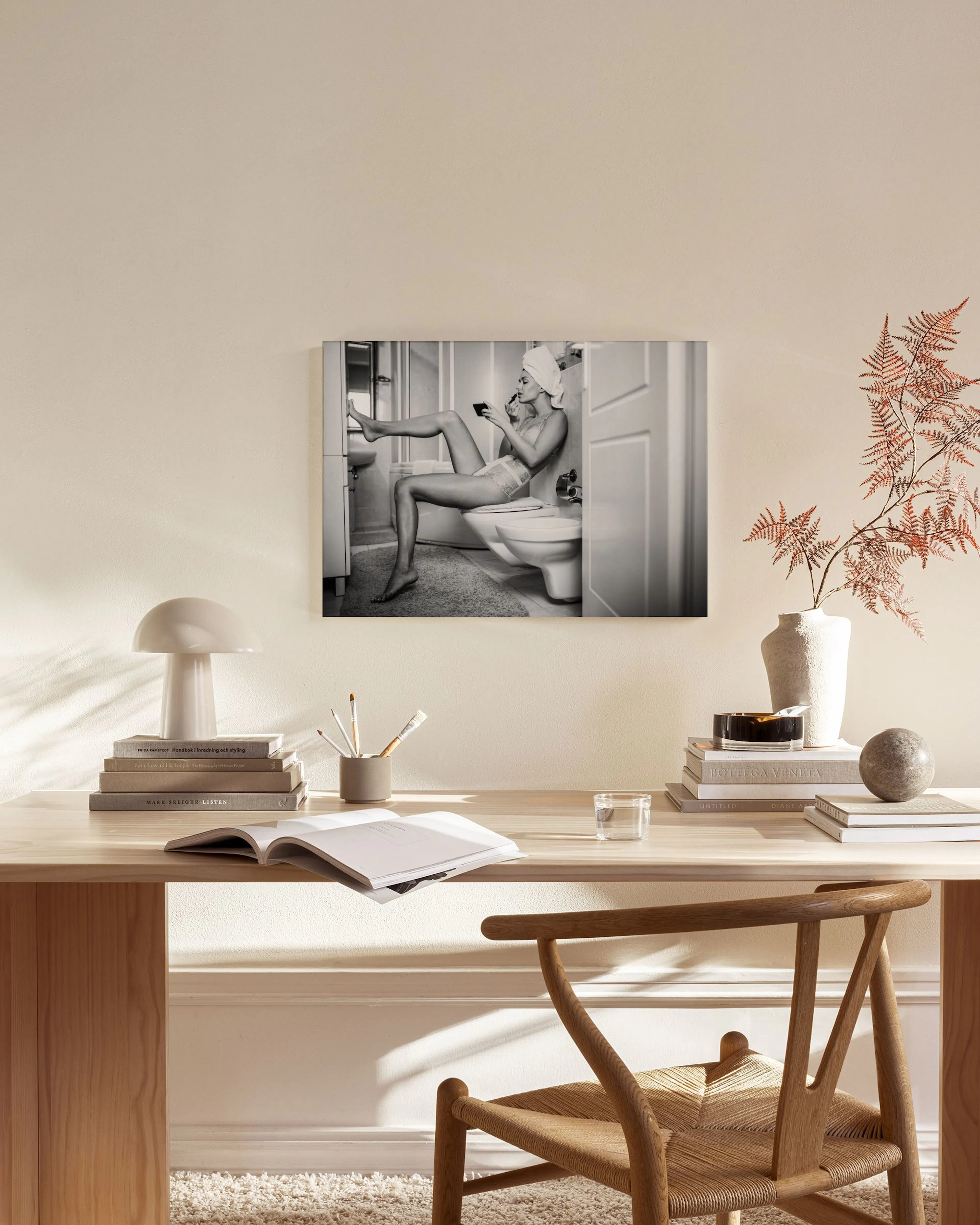 A black and white canvas print of a woman in a towel applying makeup while sitting on a toilet, hanging above a console table in