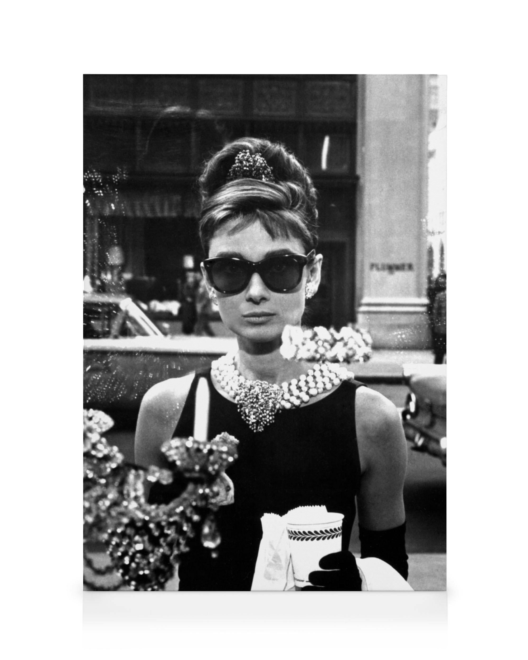 A black and white canvas print featuring Audrey Hepburn in sunglasses, a black dress, and pearl necklace, holding a cup.