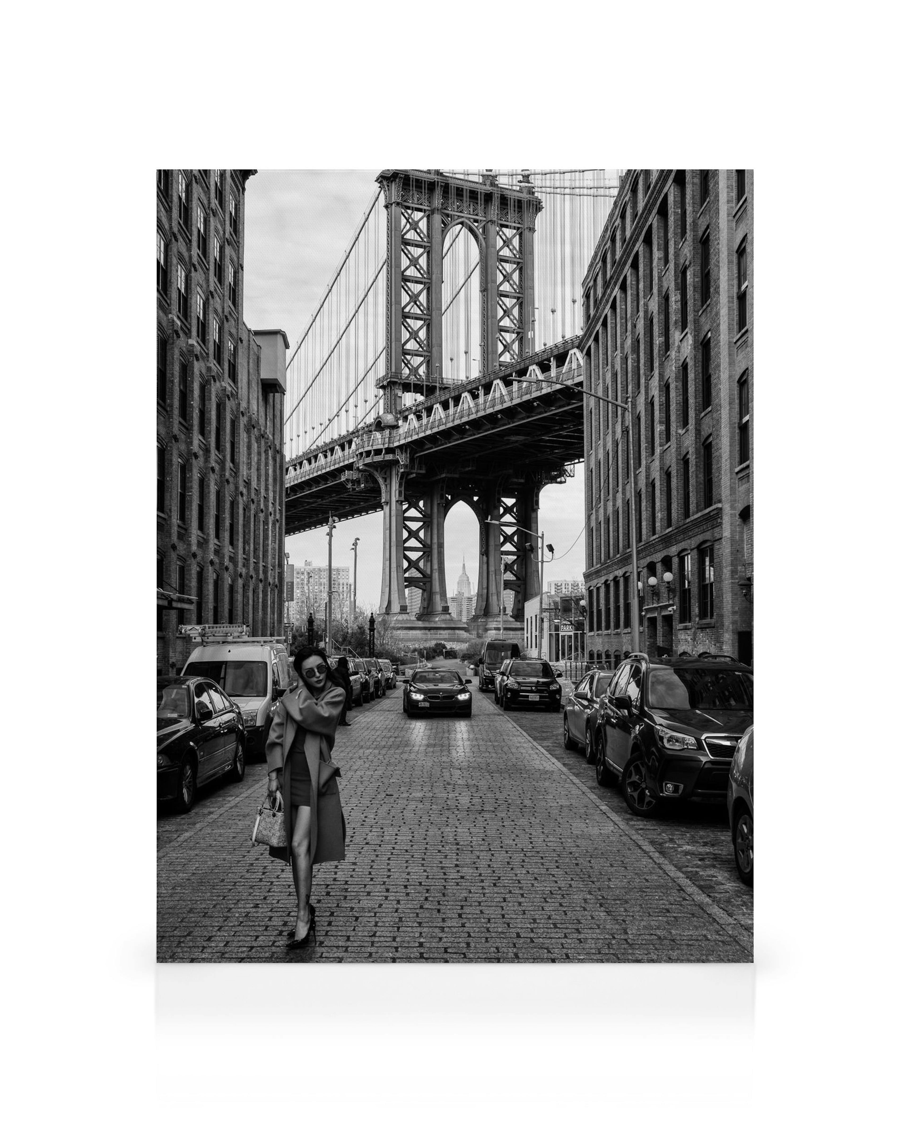 Black and white cityscape with woman walking on cobbled street, cars parked, and Manhattan Bridge overhead.