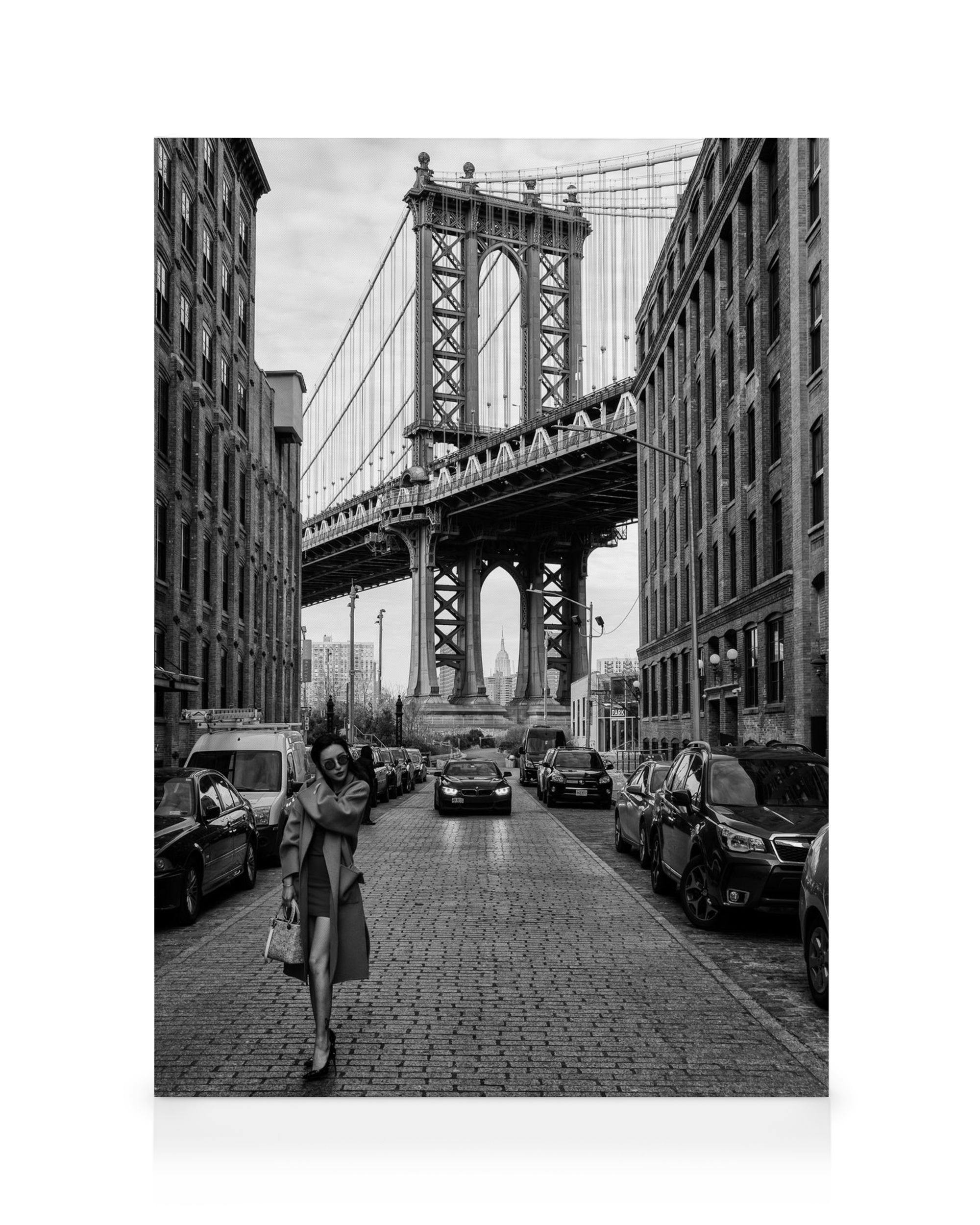 Black and white photograph of a woman in a trench coat on a cobblestone street with Manhattan Bridge in New York.