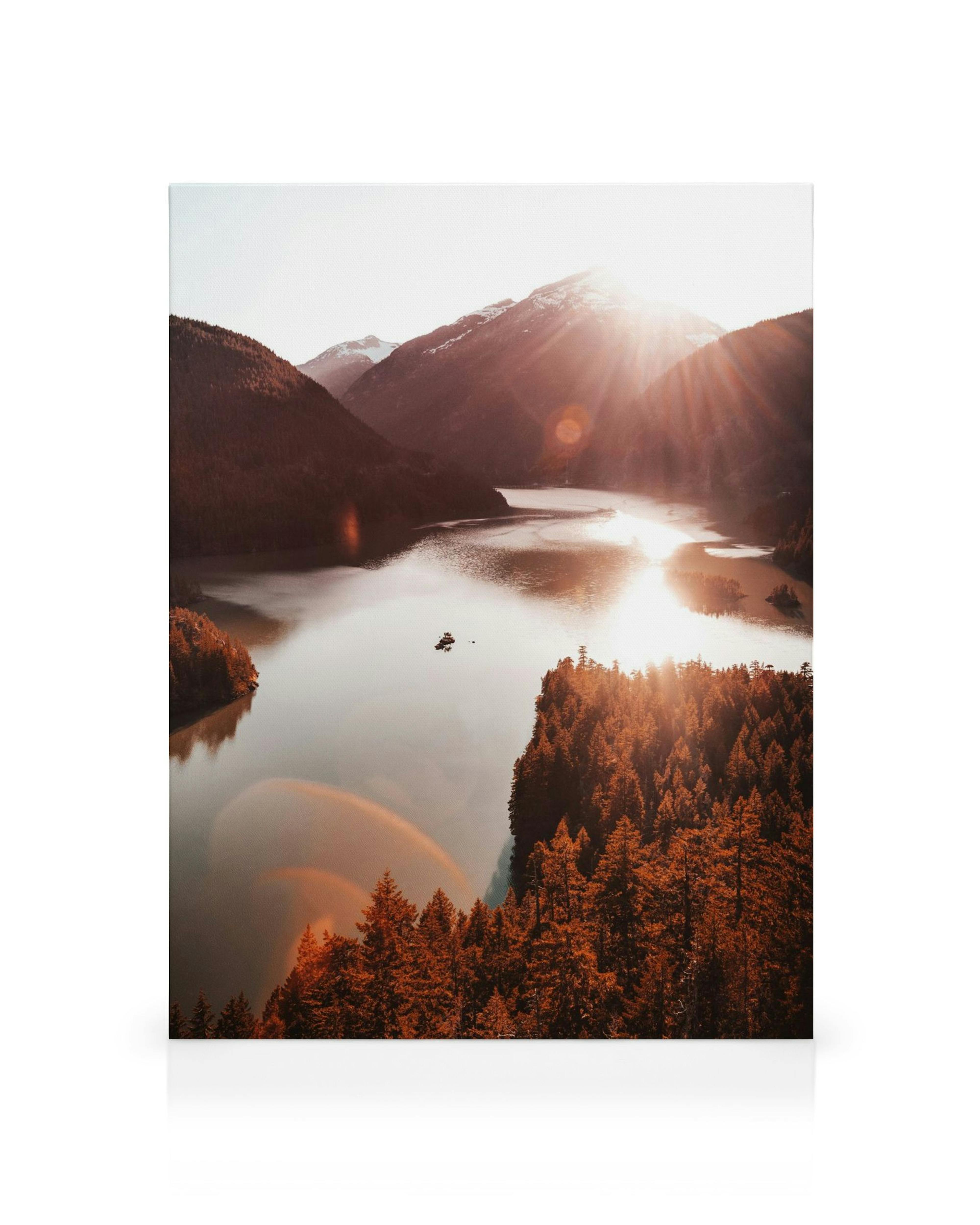 Fotografie eines Sees im Sonnenuntergang mit roten Bäumen im Vordergrund und Bergen im Hintergrund, Sonnenstrahlen reflektieren 