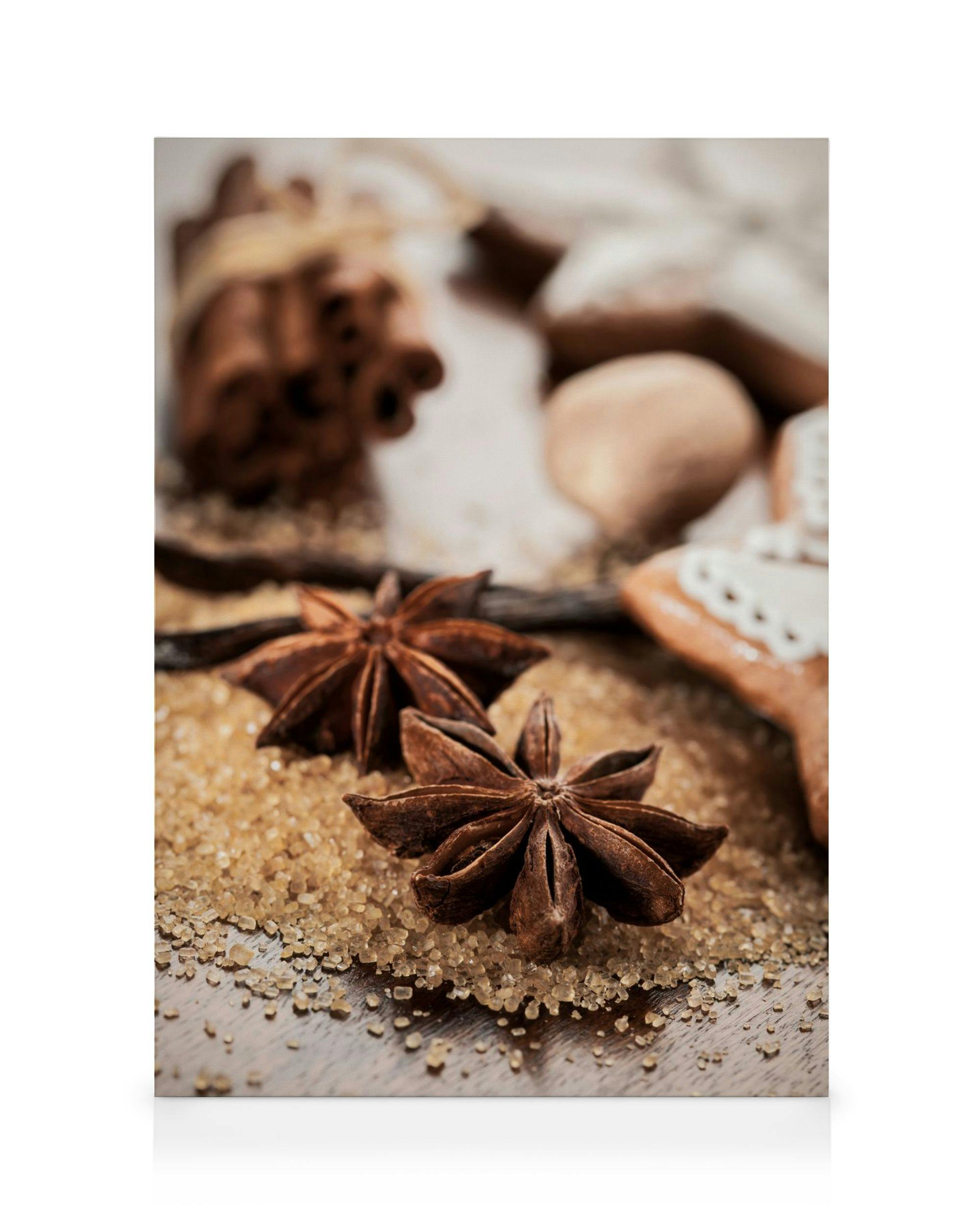 Canvastavla med närbild av stjärnanis och kanelstänger på farinsocker, med en vaniljstång och bakverk i bakgrunden.