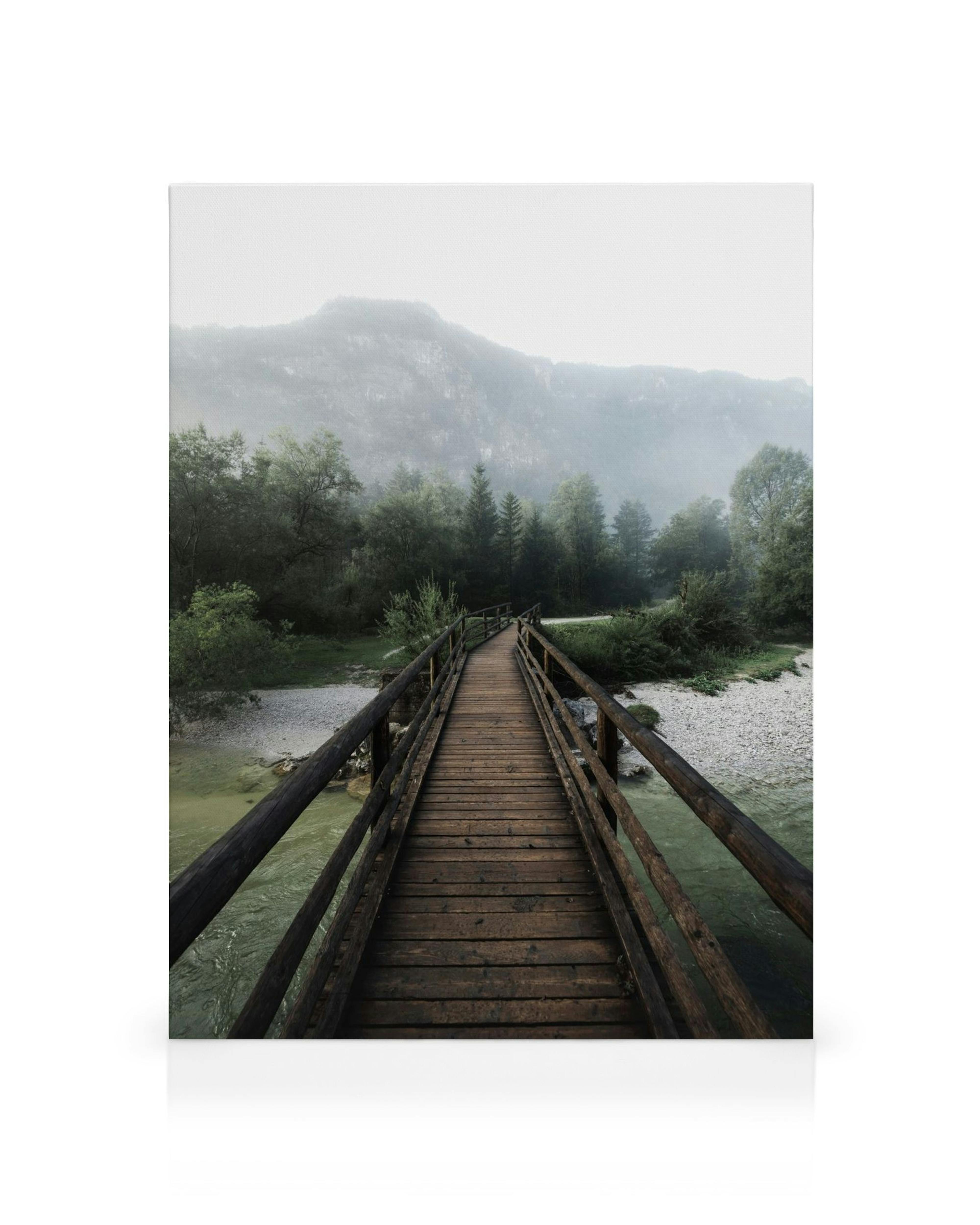 Lienzo de un puente de madera rústico sobre un río claro hacia montañas brumosas y árboles verdes bajo un cielo nublado.