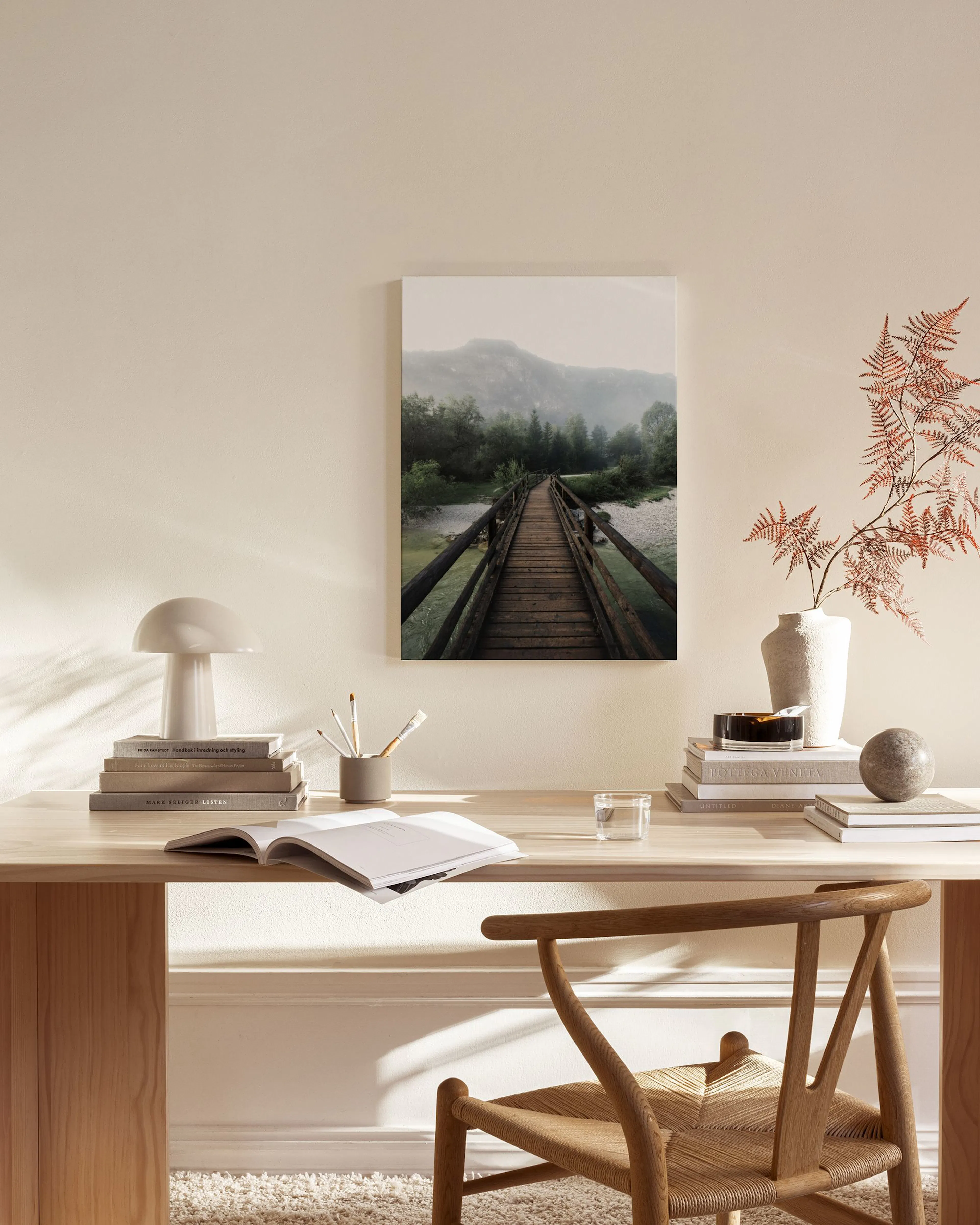 A canvas print of a wooden footbridge over water, leading towards misty mountains and evergreen trees, hanging in a minimalist l