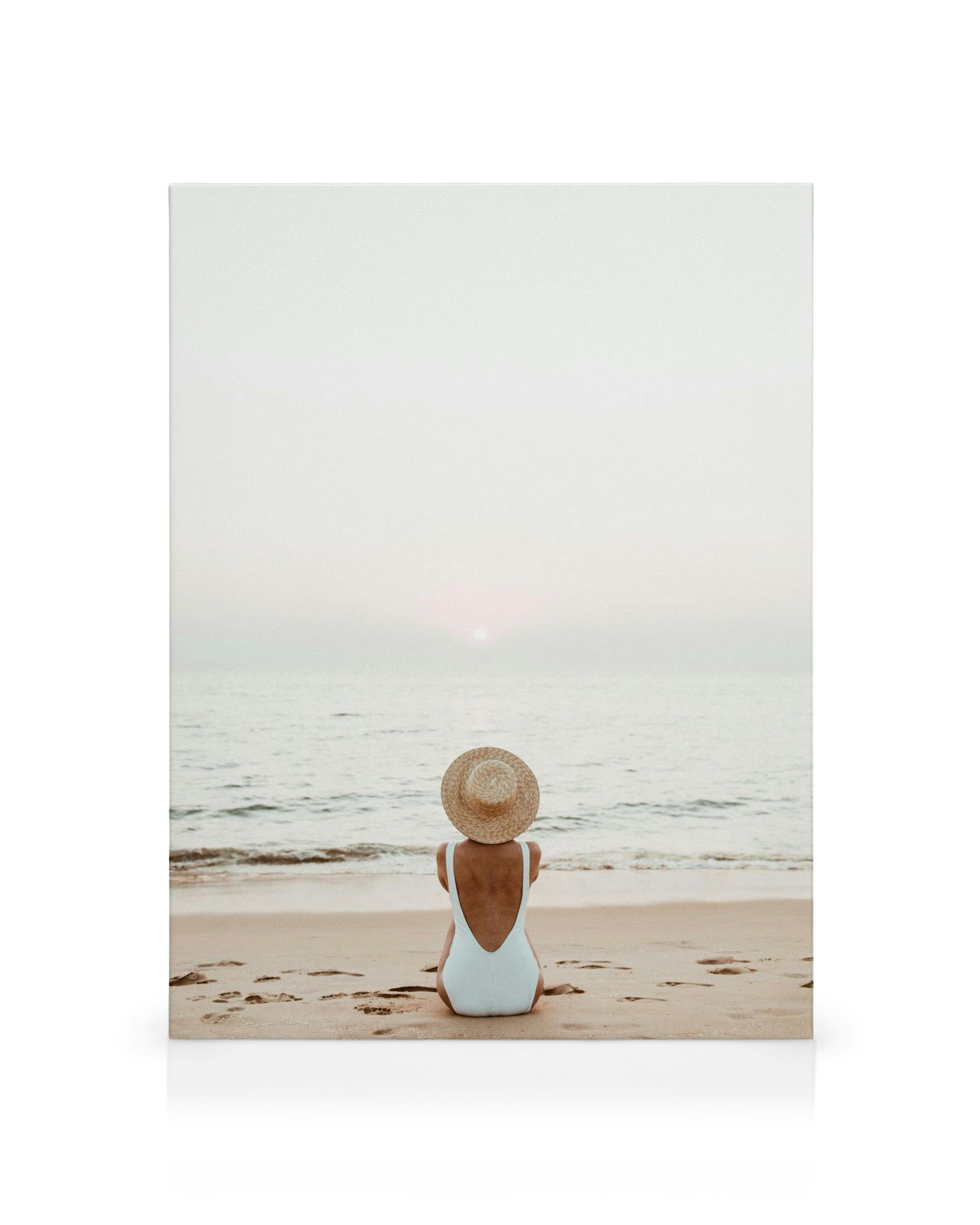 Stampa su tela, persona seduta sulla spiaggia con cappello di paglia e costume da bagno blu, mare calmo, atmosfera serena.