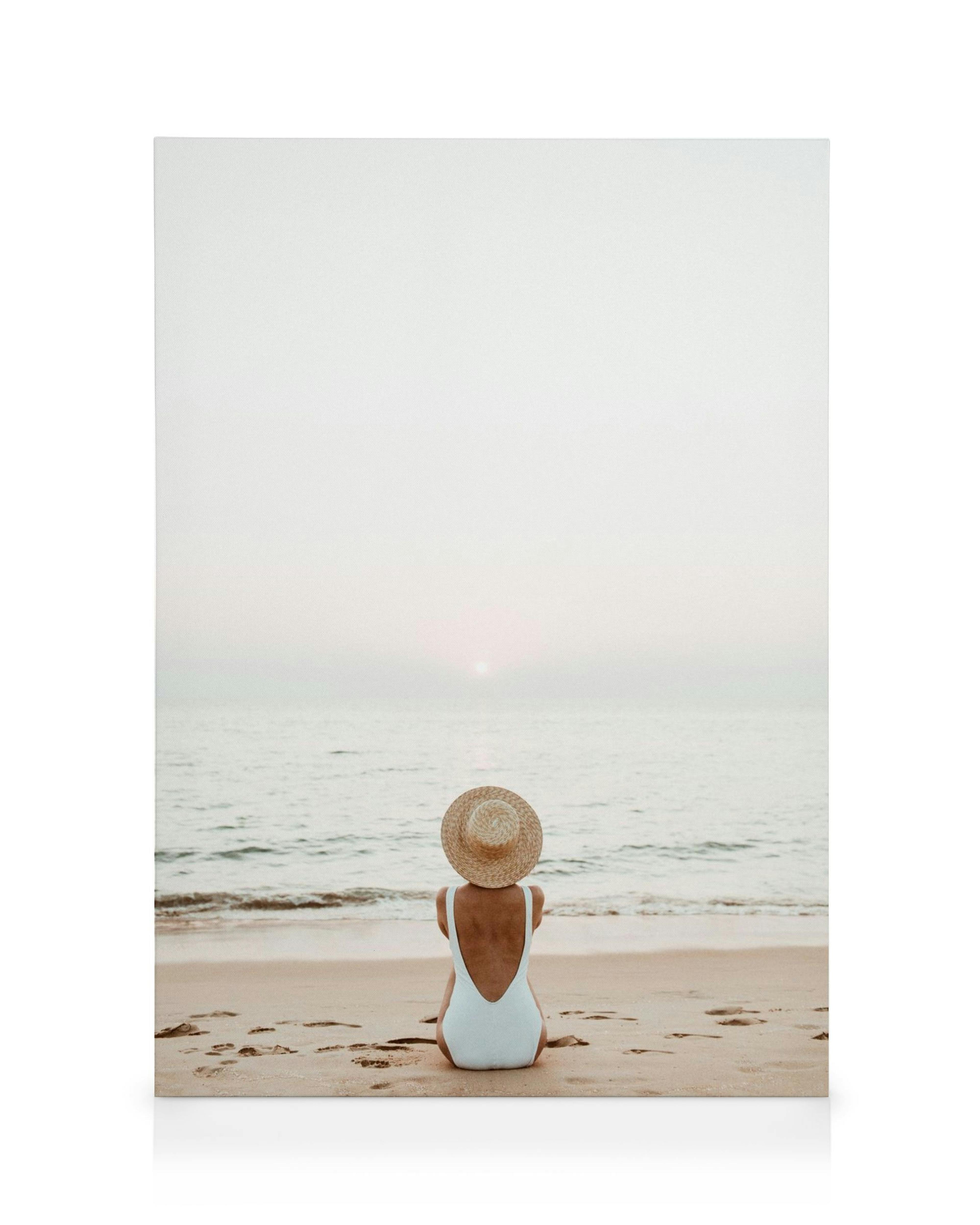 Woman in straw hat and white swimsuit sitting on a sandy beach, gazing at the ocean and sunset in a canvas print.