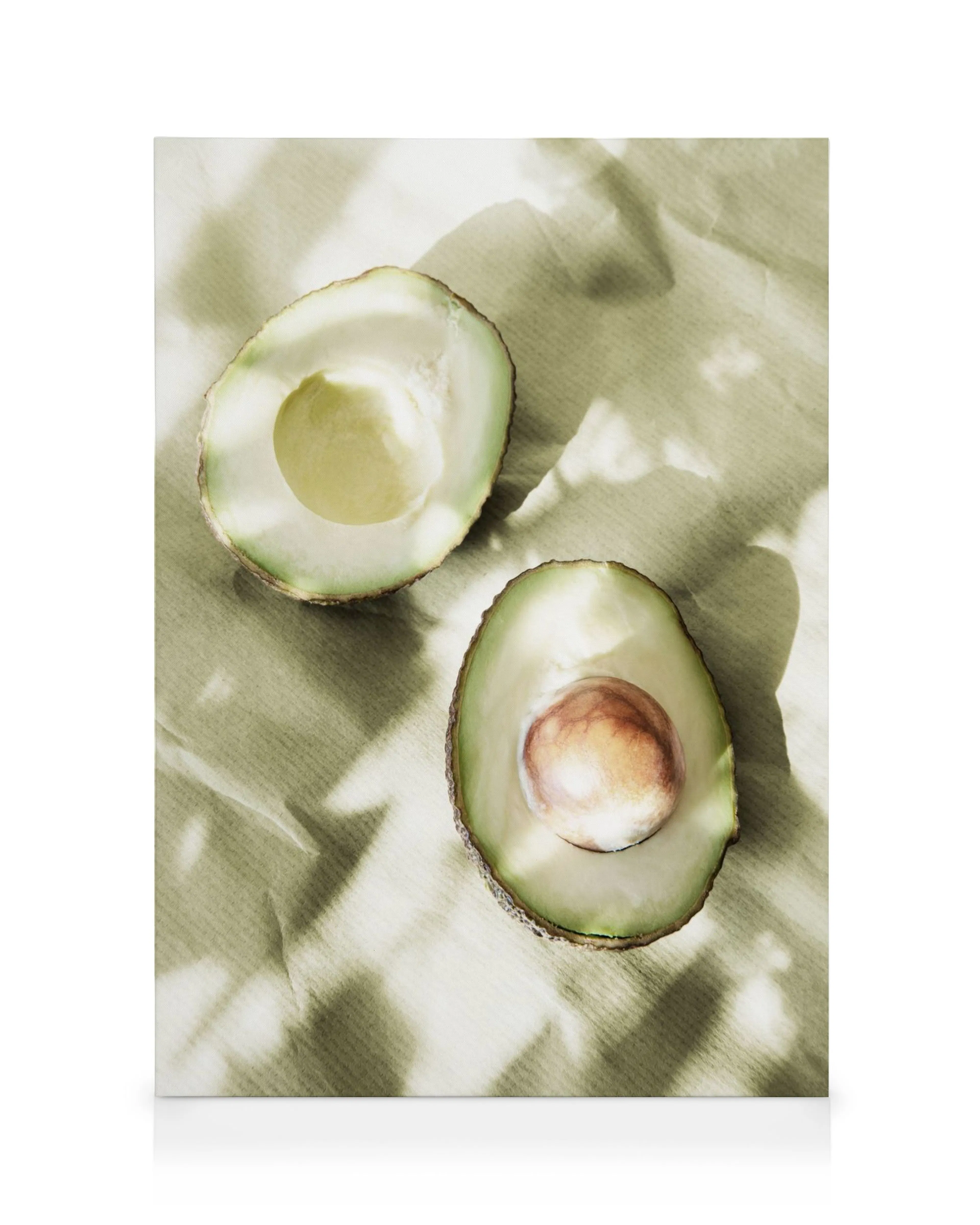 Two sunlit avocado halves, one with a pit, on a light green canvas, casting leaf shadows