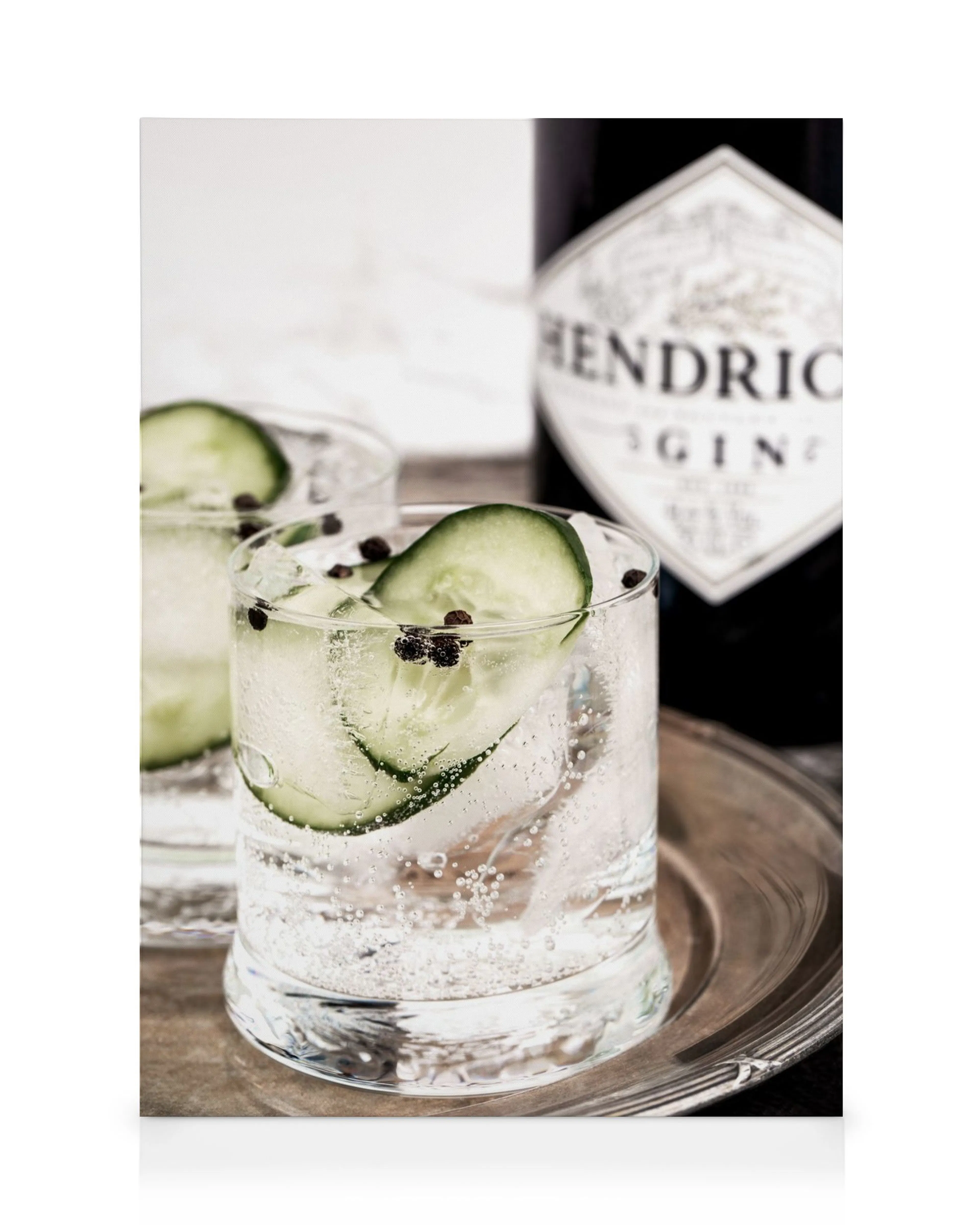 Two gin and tonic drinks with cucumber slices and peppercorns on a silver tray, with a gin bottle in the background.