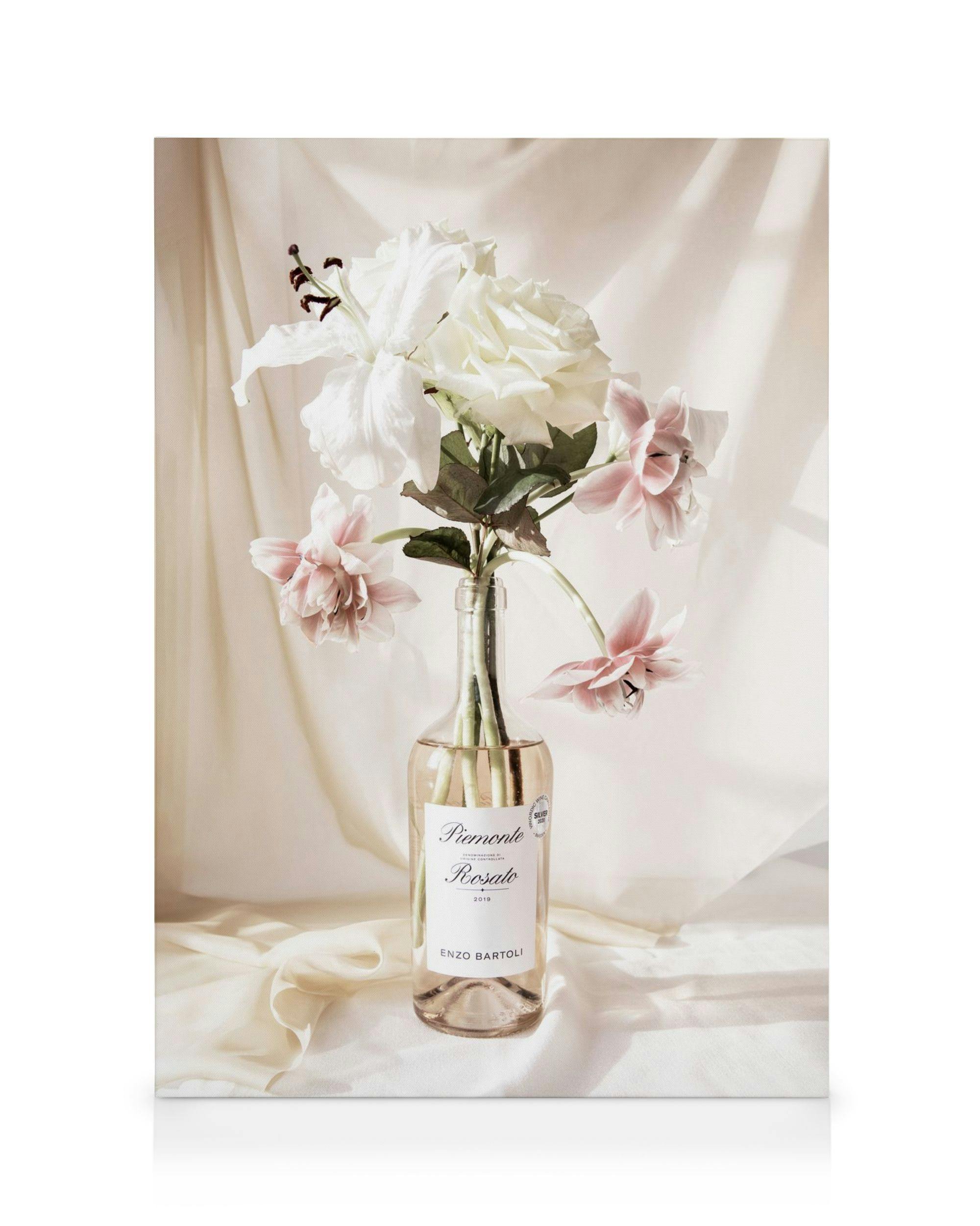 Bodegón floral con rosas y lirios blancos y rosados en una botella de vidrio sobre un paño beige.