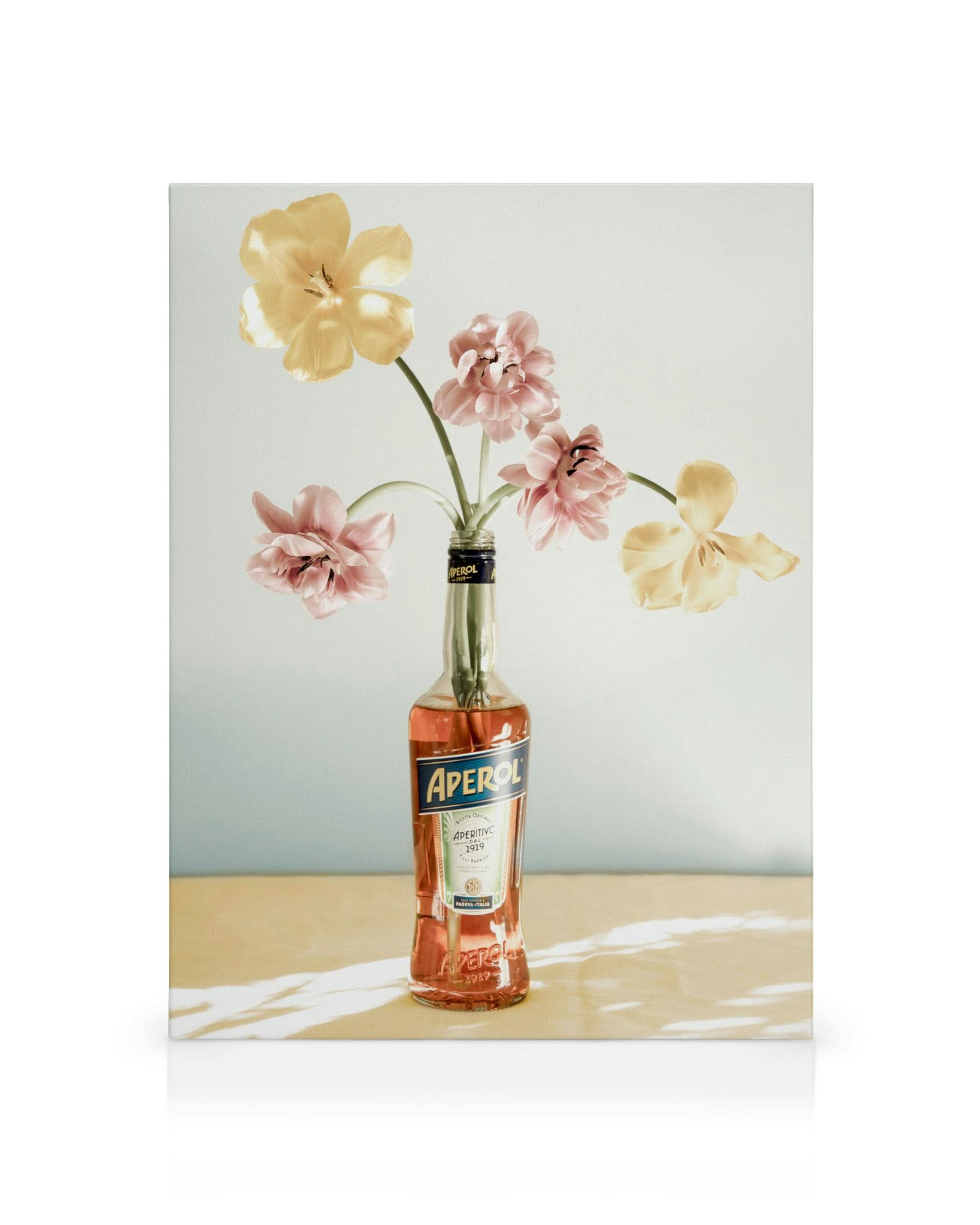 Aperol fles met roze en gele bloemen, werpt bladvormige schaduw op lichte achtergrond. Decoratieve kunst.