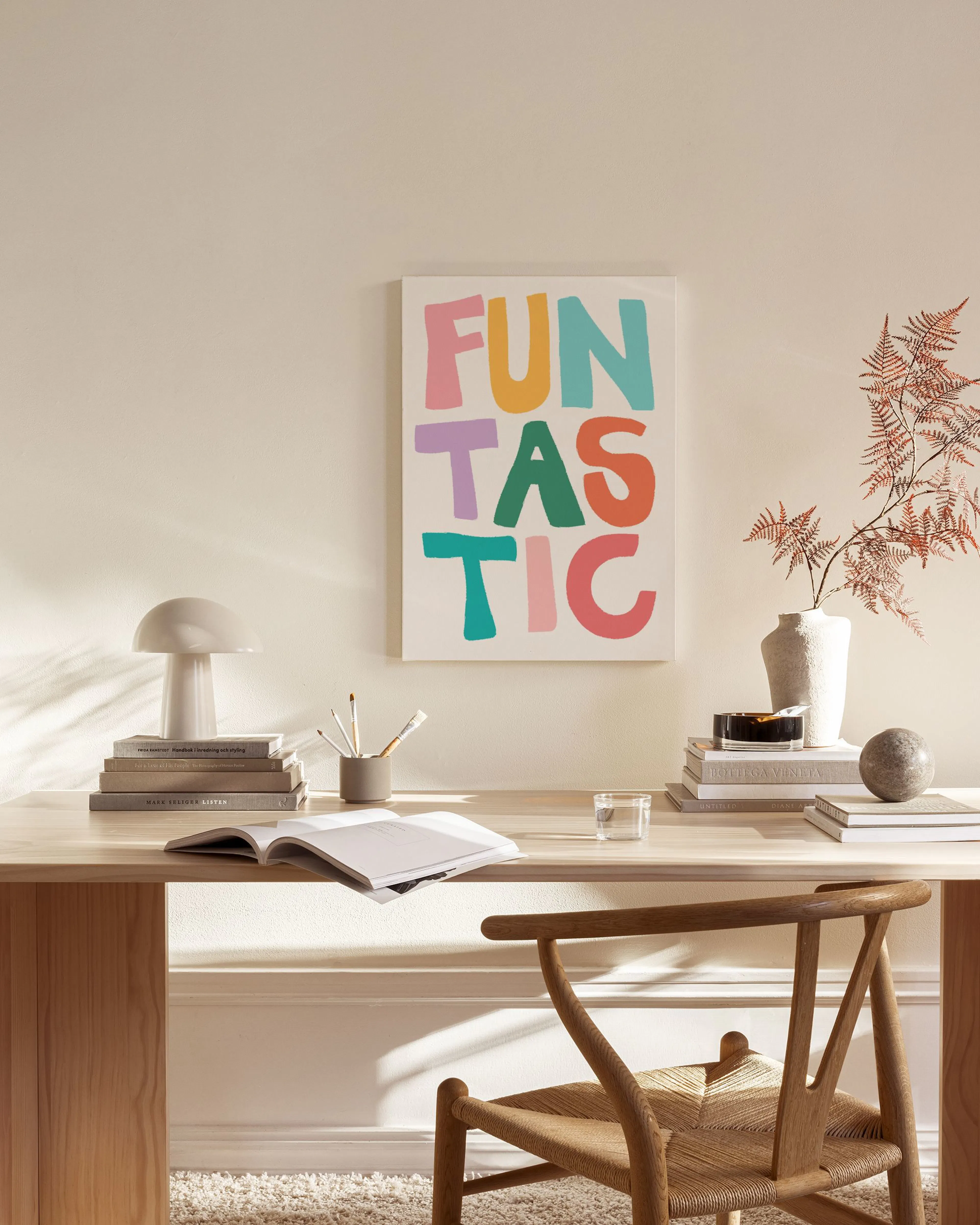 Tela decorativa com a palavra FUNTASTIC em letras coloridas, pendurada numa sala de estar com poltrona de madeira e mesa de cent