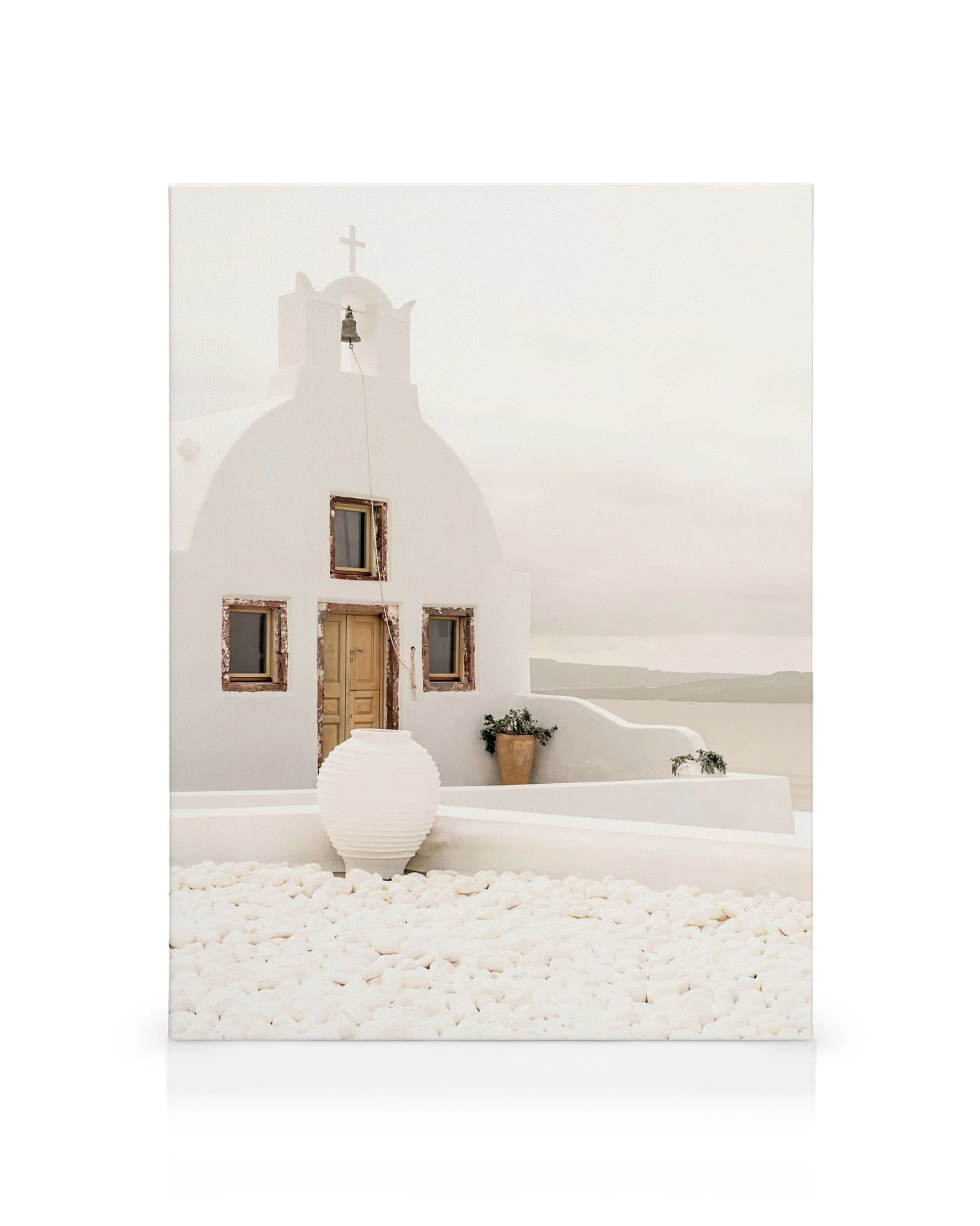 Weißes Santorini-Gebäude mit geschwungenem Dach, Holztür, Fenstern und Glocke. Weiße Kieselsteine und Pflanzen.