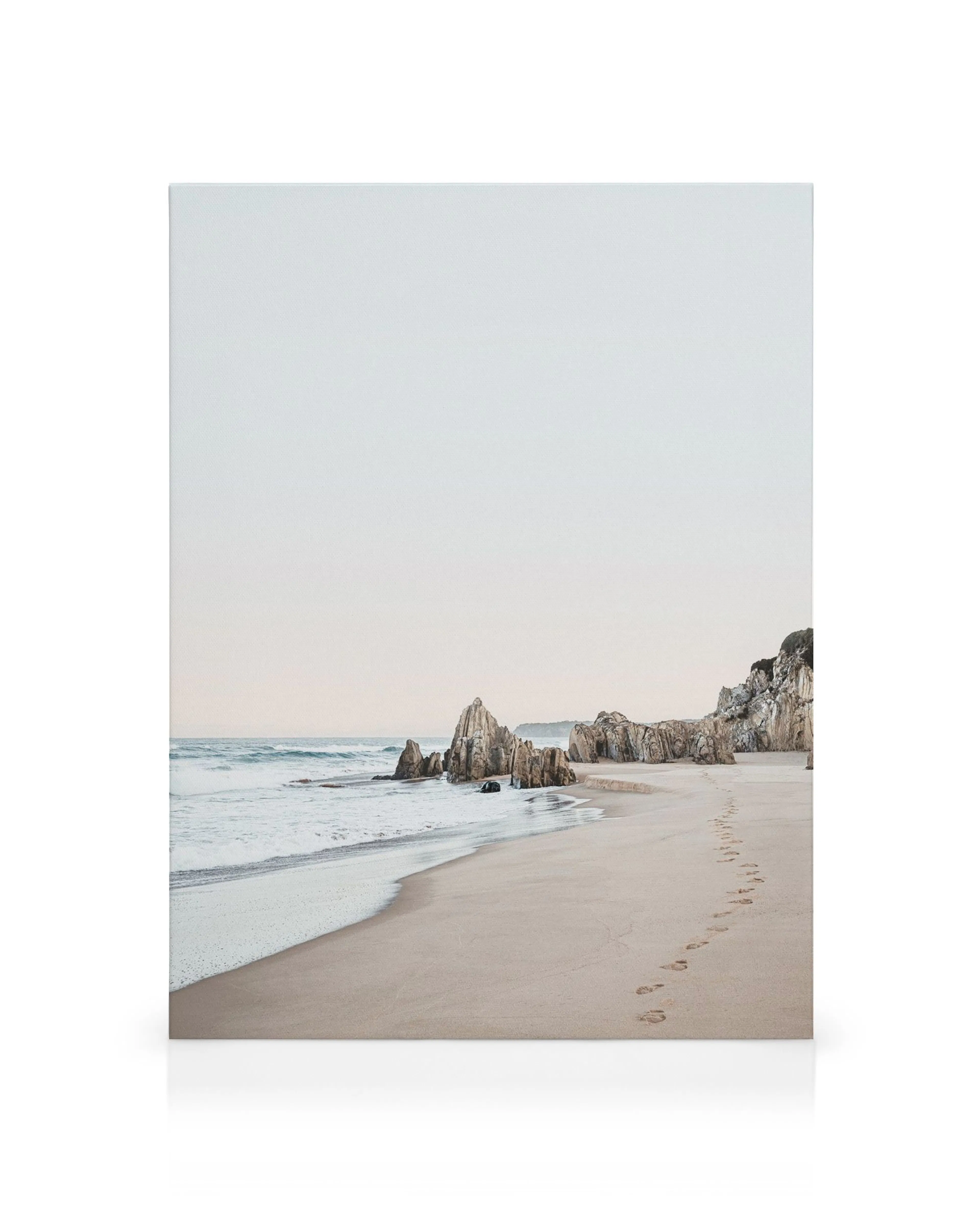 Footprints on a sandy beach leading to rocks and the sea, with muted blue, brown, and beige tones.