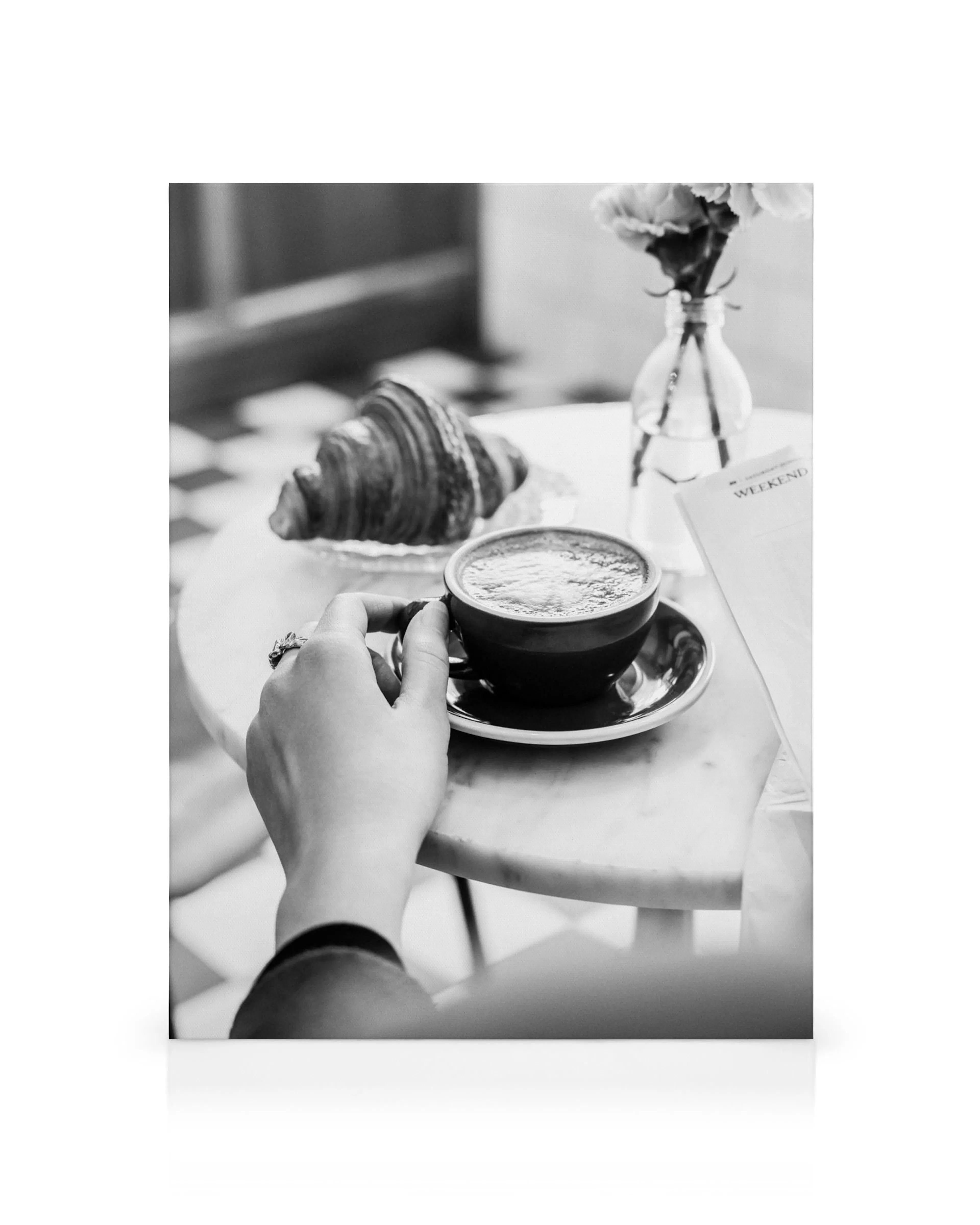 Zwart-wit canvas van hand die koffiekopje vasthoudt met croissant en bloemenvaas op tafel.