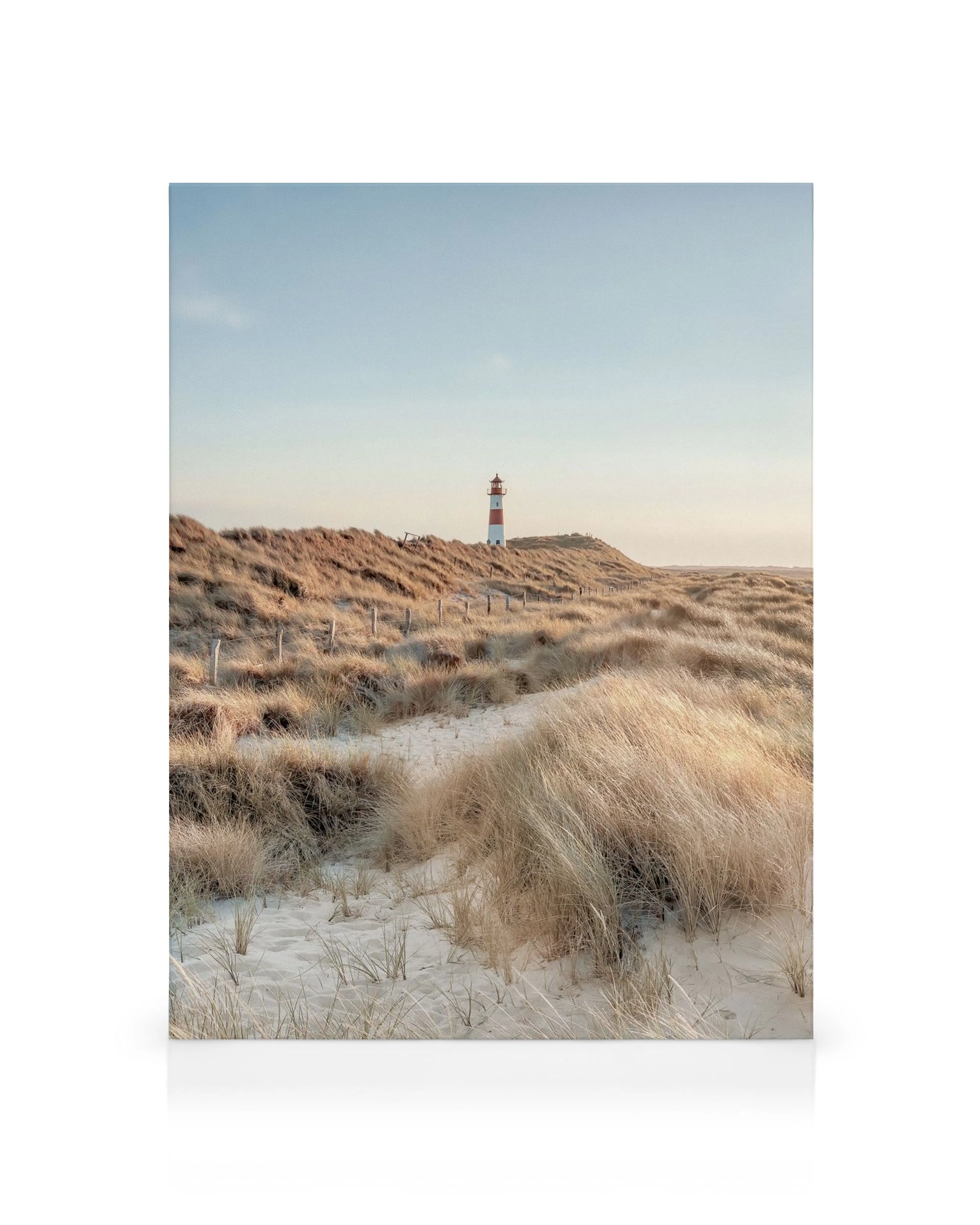 Canvas print of a sandy path winding through grass dunes to a distant striped lighthouse under a soft blue sky.