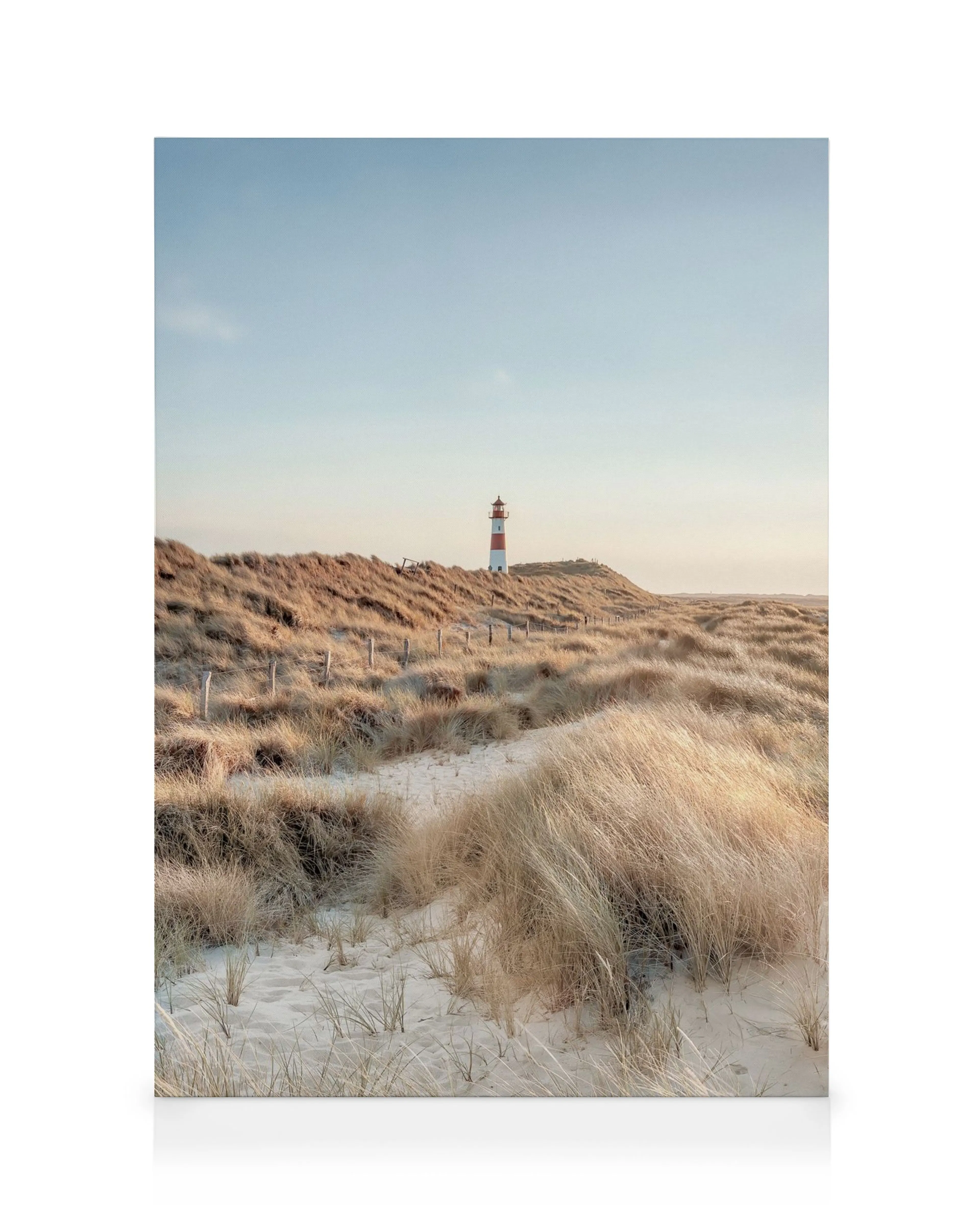 Zandpad naar gestreepte vuurtoren tussen duinen met hoog gras onder een blauwe lucht, kusttafereel.
