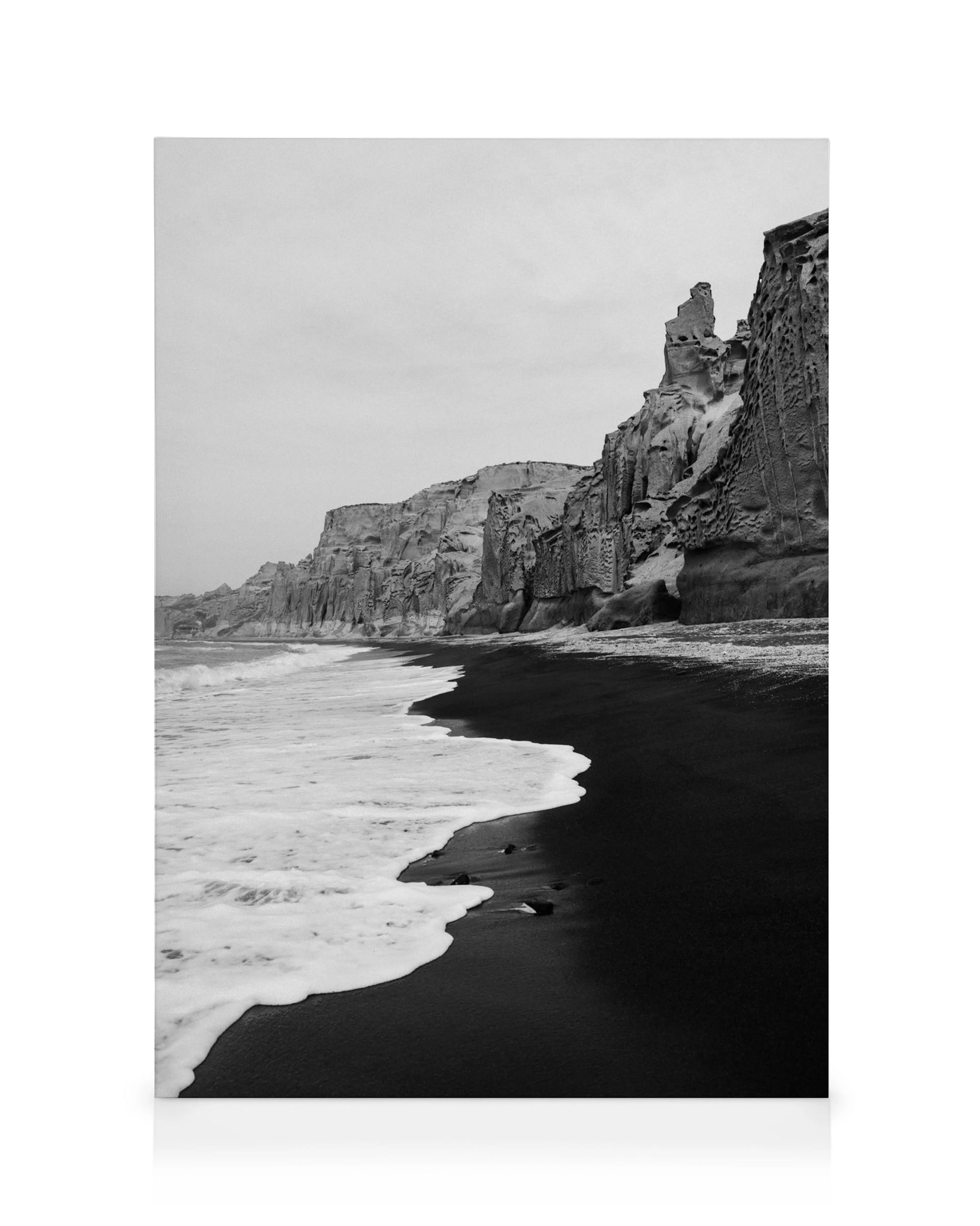 Lienzo de playa de arena negra, acantilados rocosos y olas espumosas en blanco y negro.