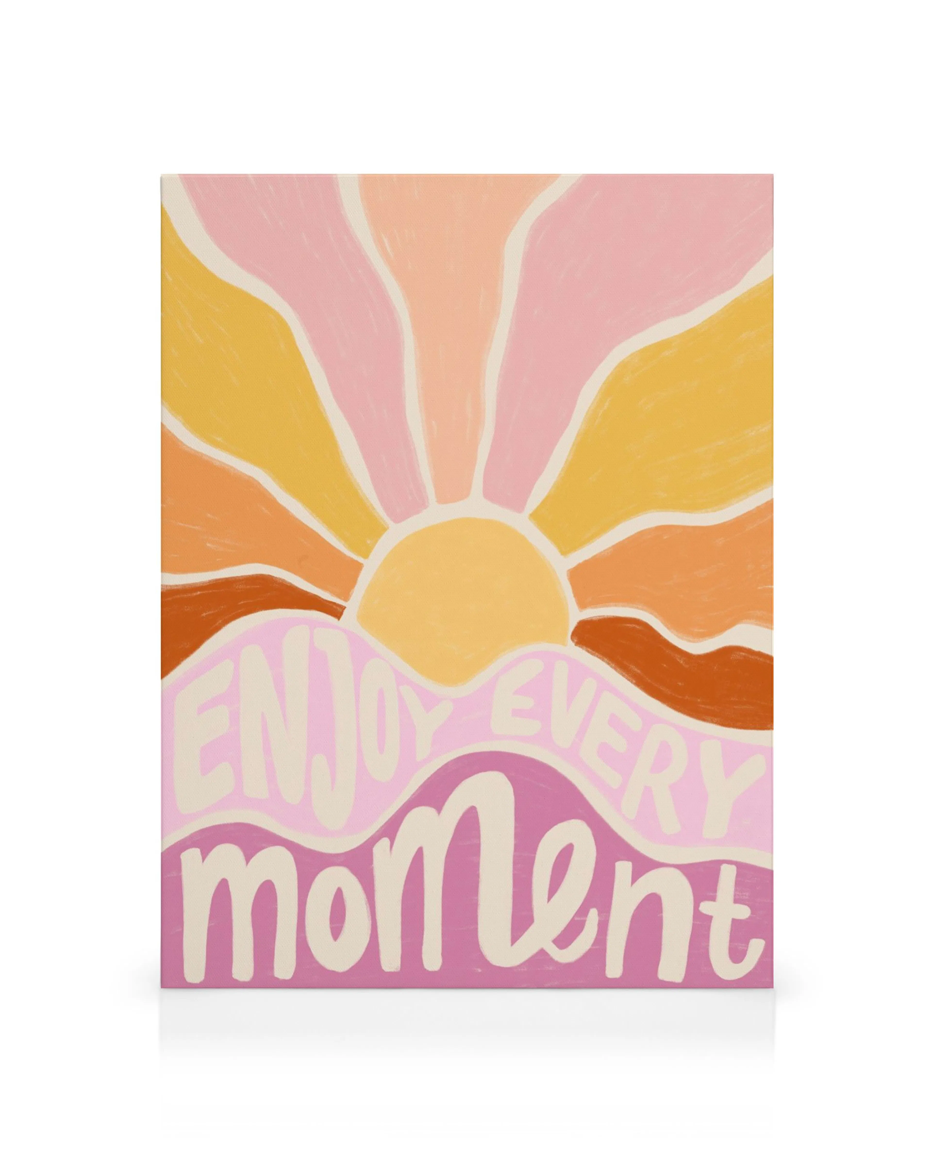 A canvas print featuring a vibrant sunset with orange, pink, and yellow rays over wavy pink hills, with Enjoy Every Moment text.