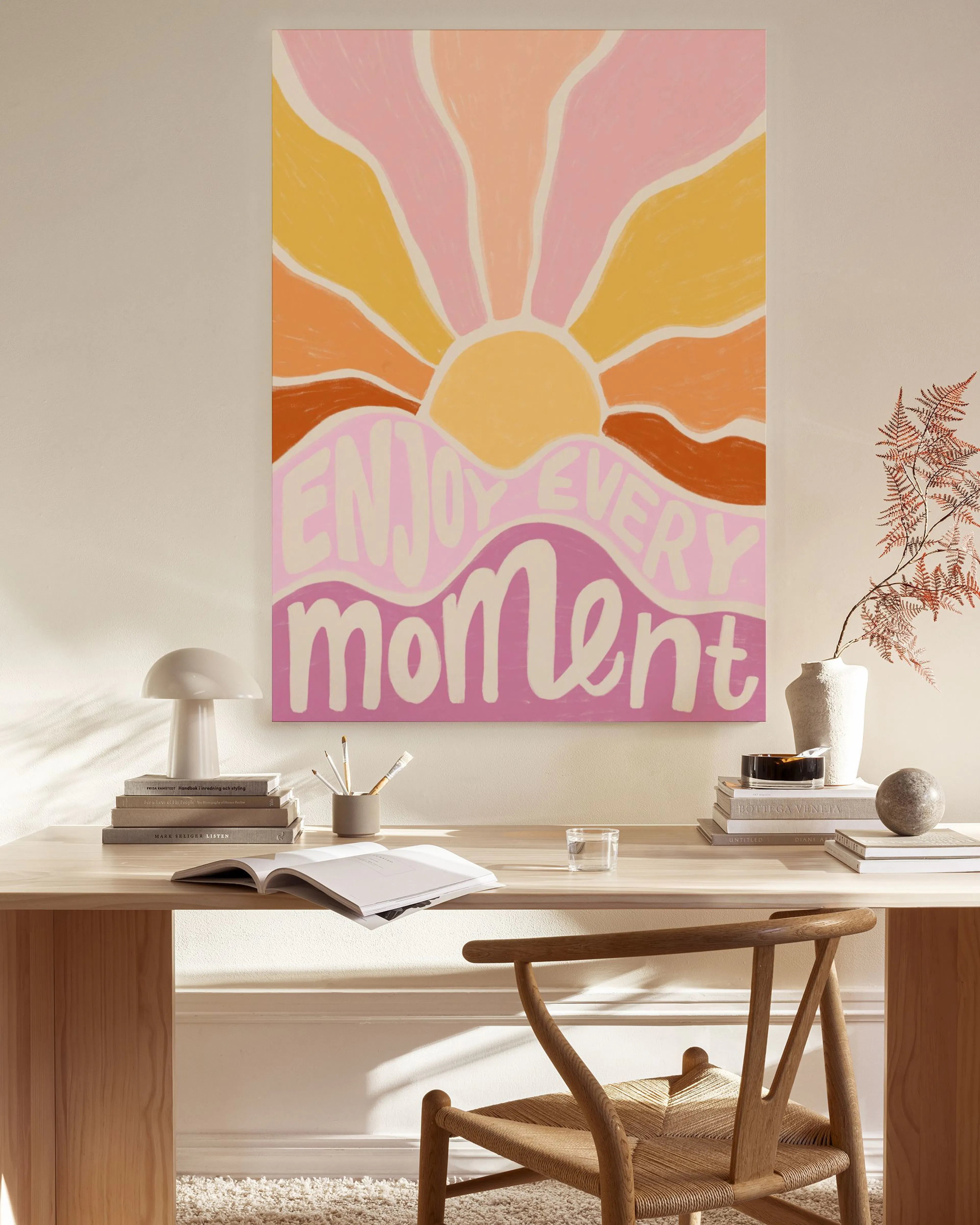 A vibrant canvas print with a sunburst in shades of pink, yellow, and orange, above text reading Enjoy Every Moment.