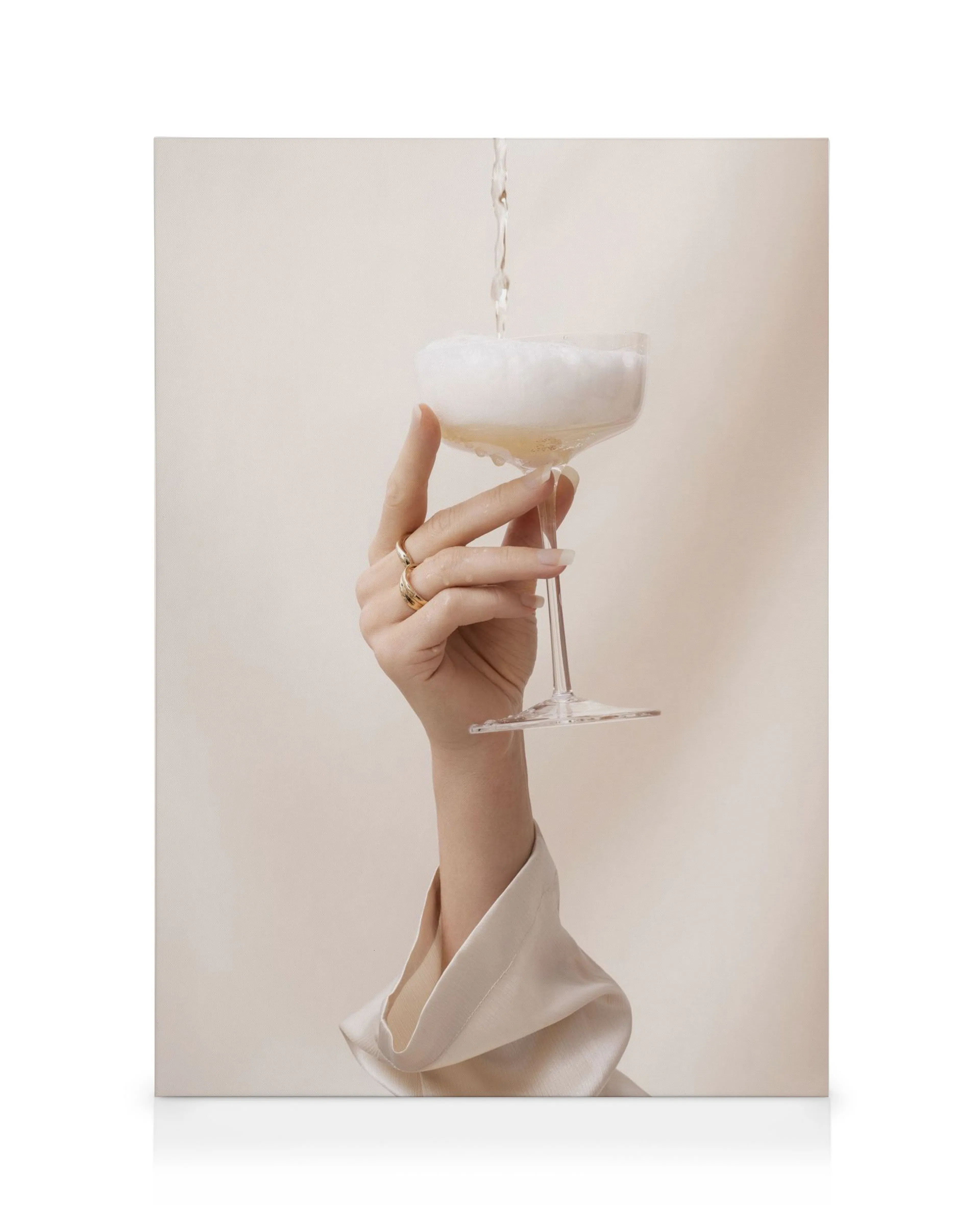 Hand hält gefülltes Champagnerglas, goldene Ringe, beige Ärmel, minimalistischer Stil