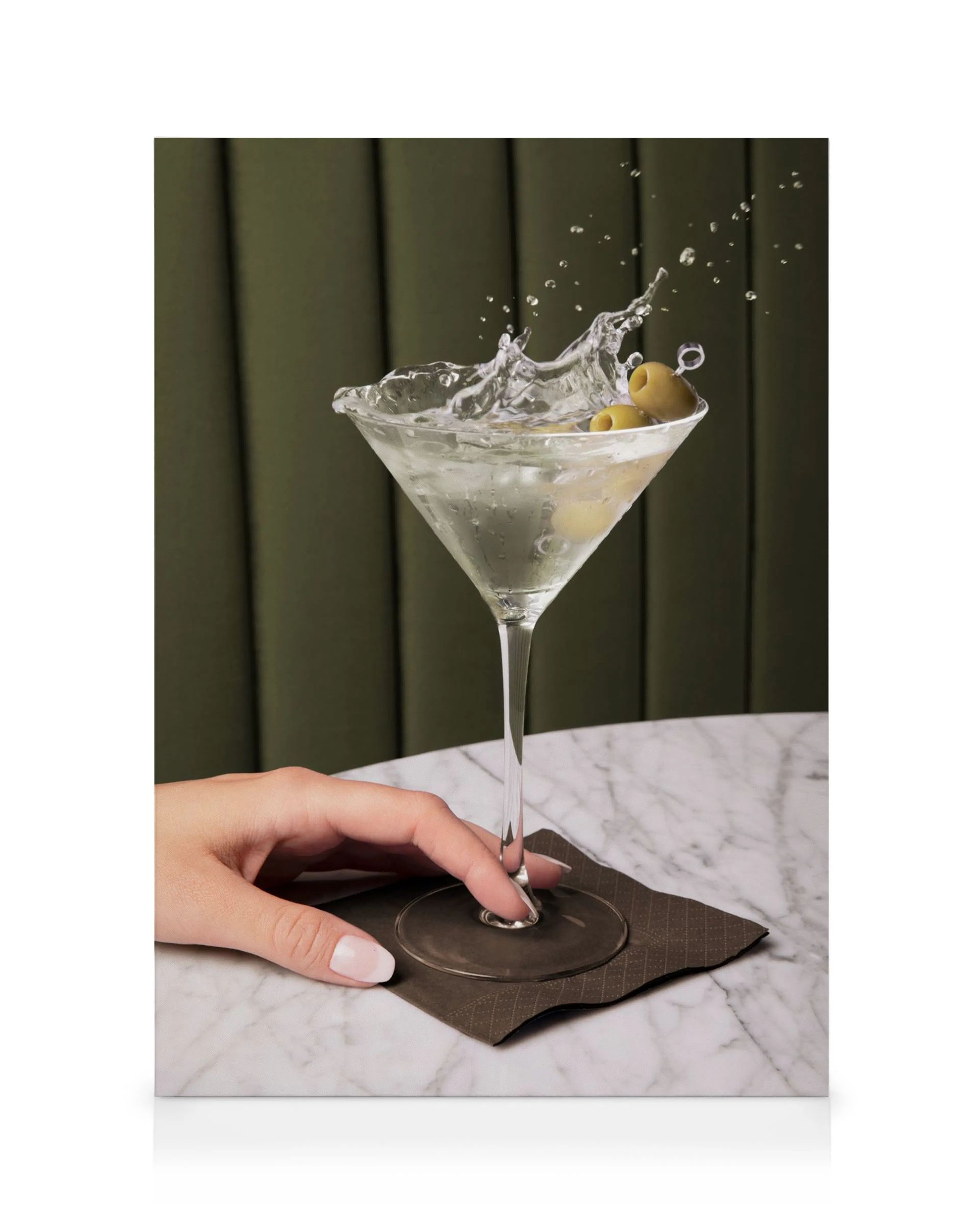 Leinwanddruck eines Martini-Glases mit spritzender Flüssigkeit und Oliven, gehalten von einer Hand, auf Marmortisch vor grünem H
