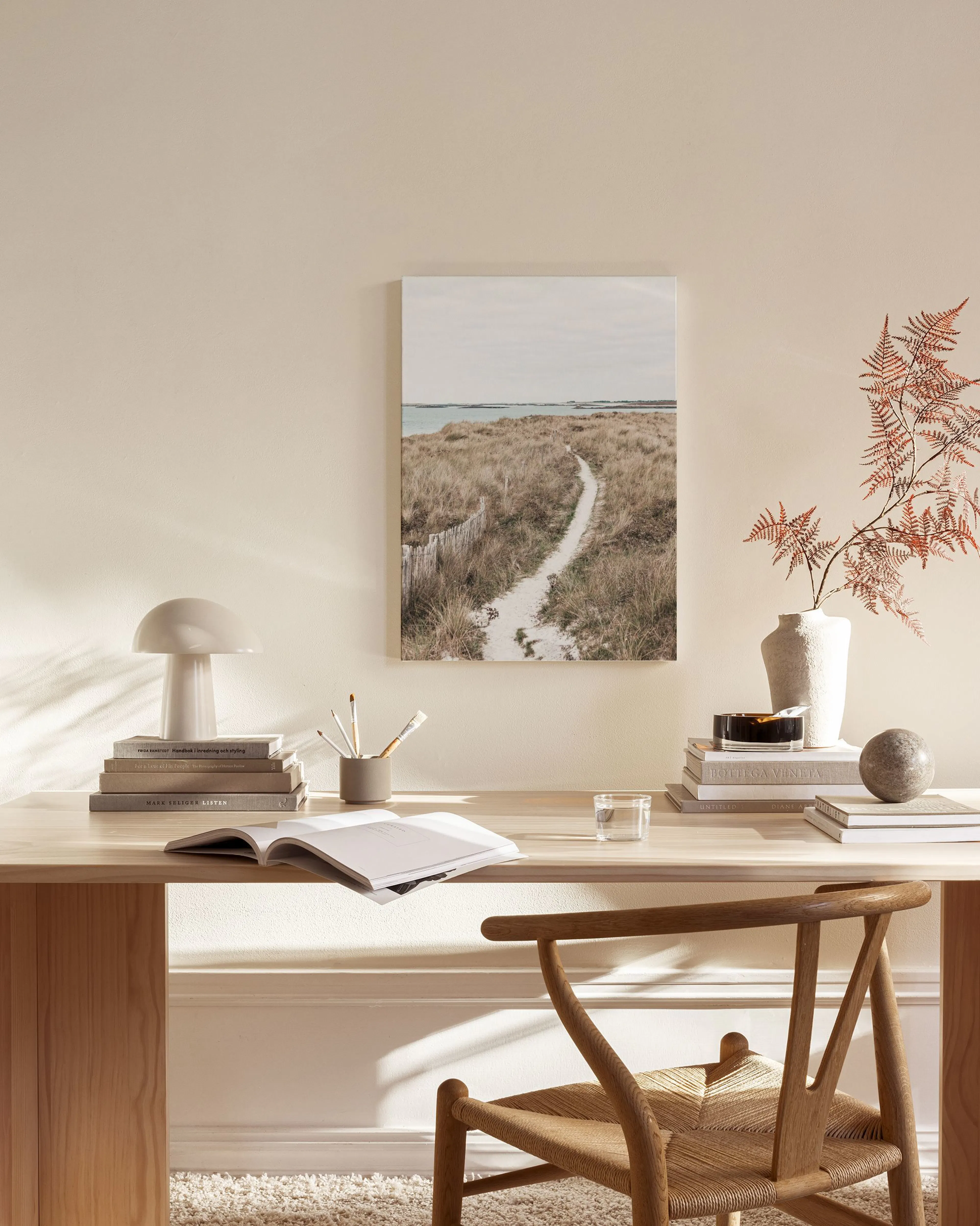 Leinwandbild einer Dünenlandschaft mit Pfad und Meer, über einem Beistelltisch mit Lampe und Büchern im Wohnzimmer.
