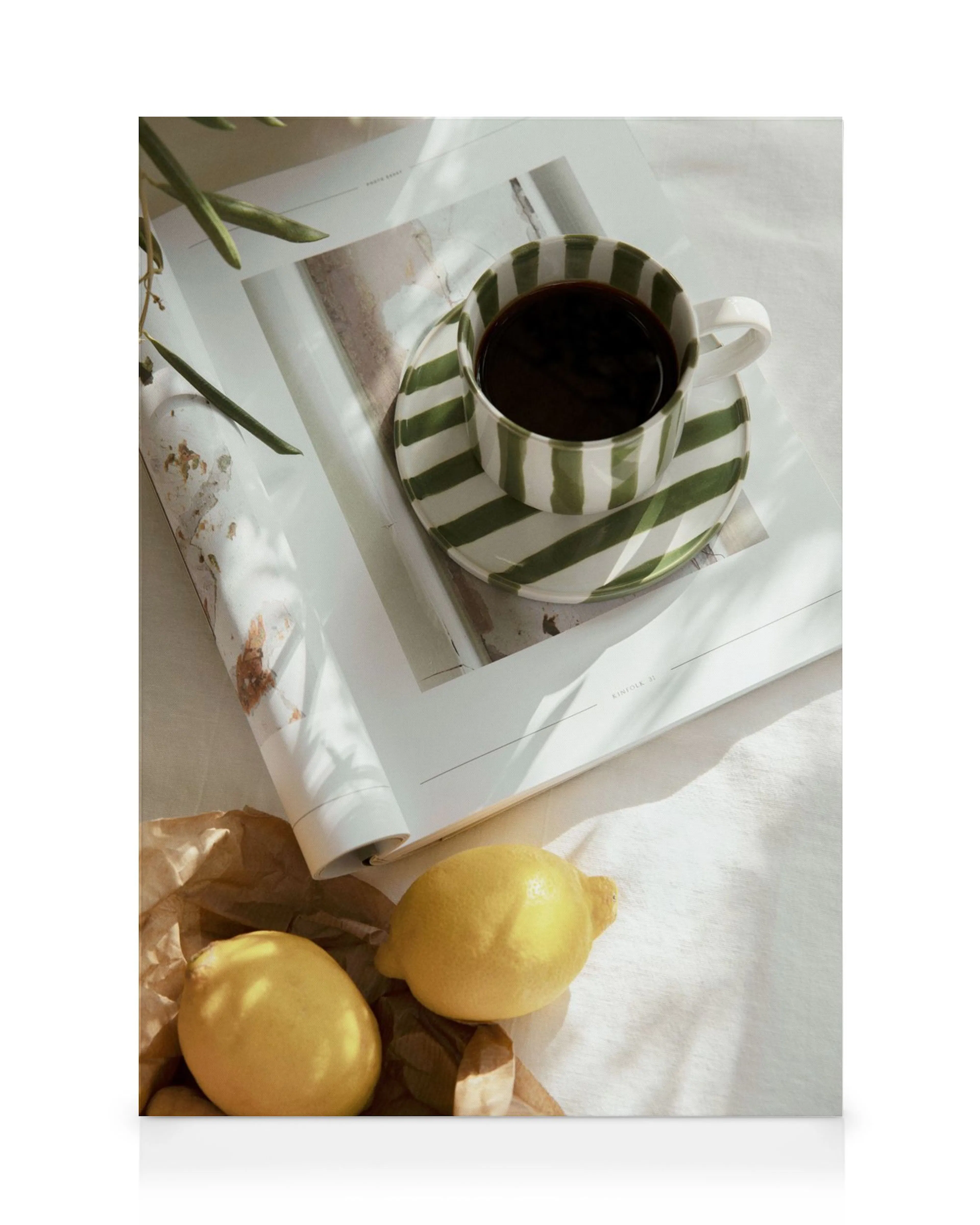 Un lienzo con una taza de café a rayas verdes y blancas sobre una revista, con limones y papel debajo.