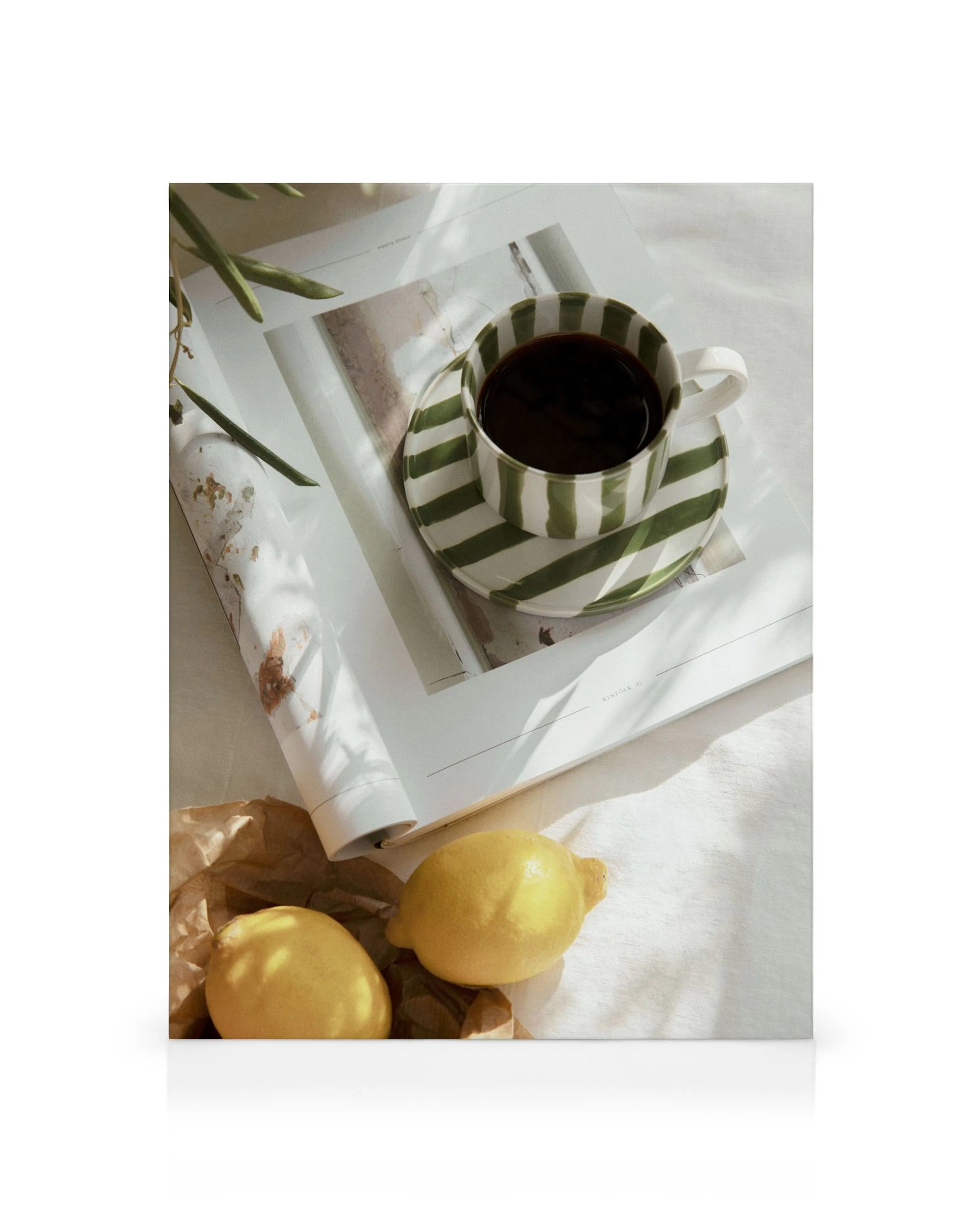 A canvas print of a green striped coffee cup on an open magazine with two lemons and a brown paper bag.