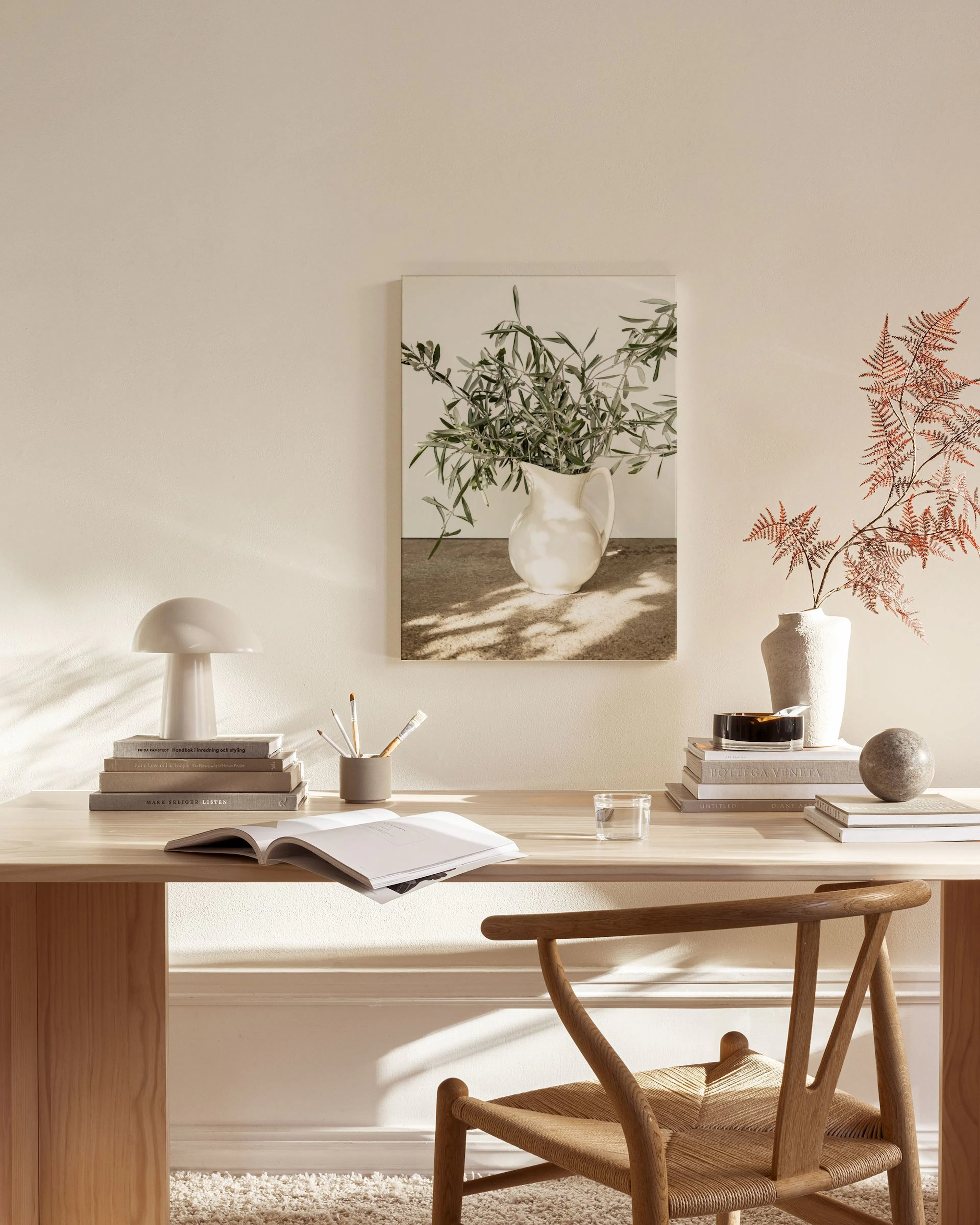 A canvas print of olive branches in a white pitcher with dappled sunlight, hanging in a minimalist living room above a wooden co