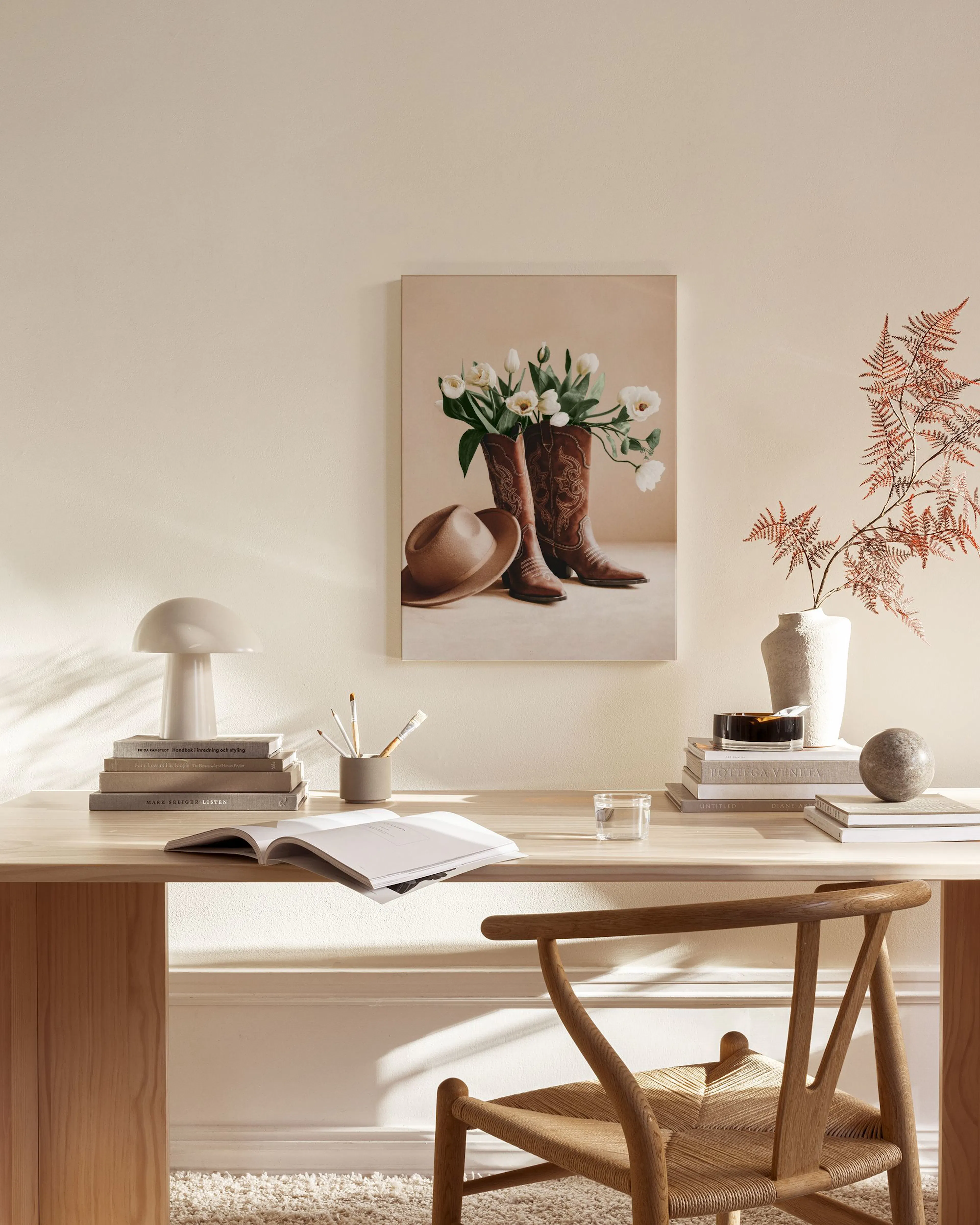 Canvastavla med bruna cowboyboots fyllda med vita tulpaner och en hatt, hängandes i ett ljust vardagsrum.