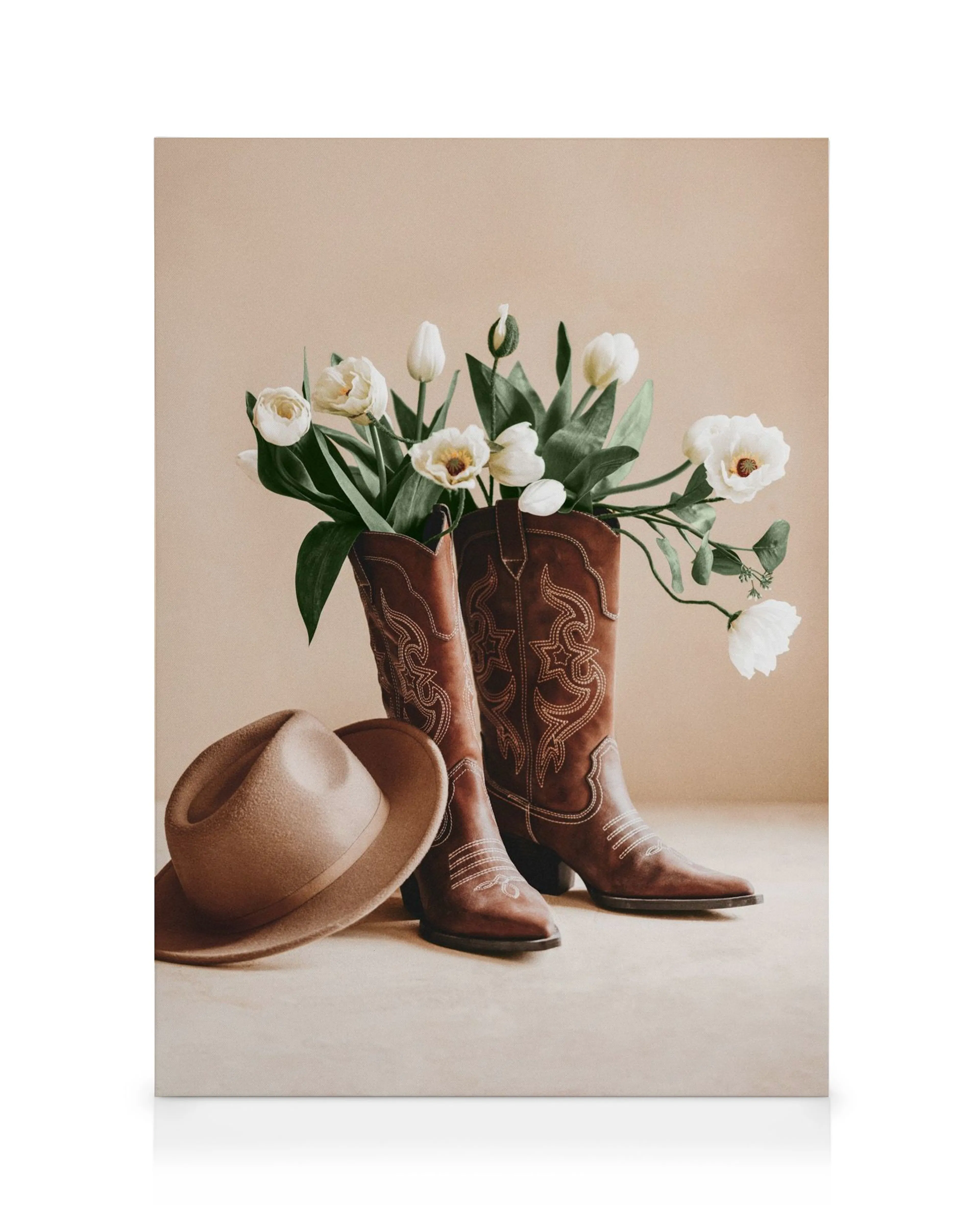 Bruna cowboyboots med vita vallmo och tulpaner, samt en brun hatt, mot en beige bakgrund. Blomstermotiv i lantlig stil.
